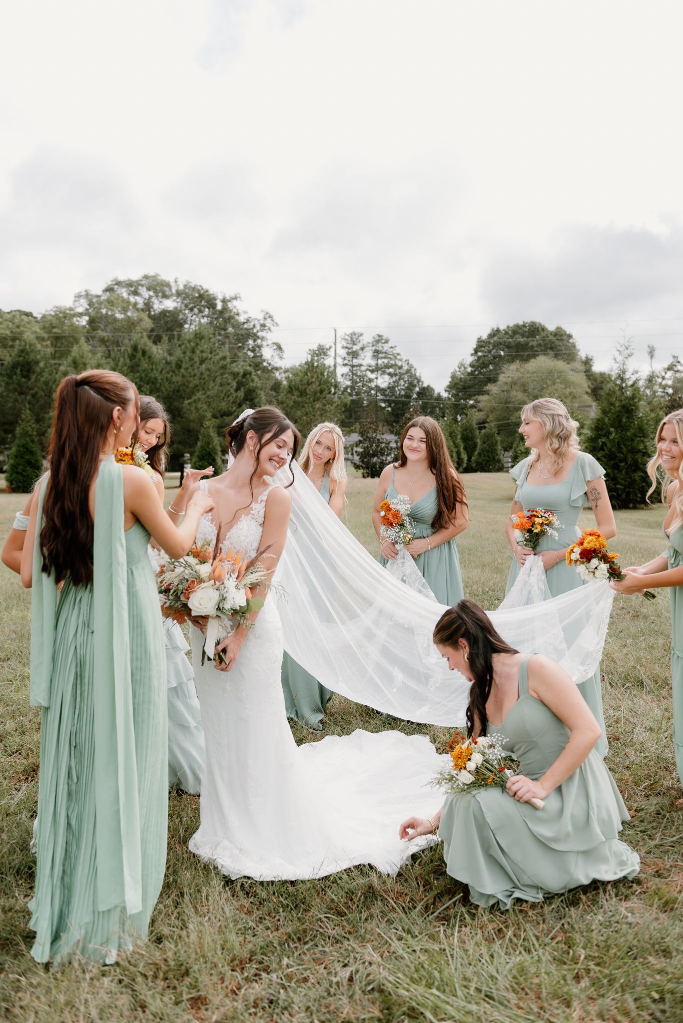 Bridesmaids in mint green dresses help arrange the bride's flowing white gown in a field.