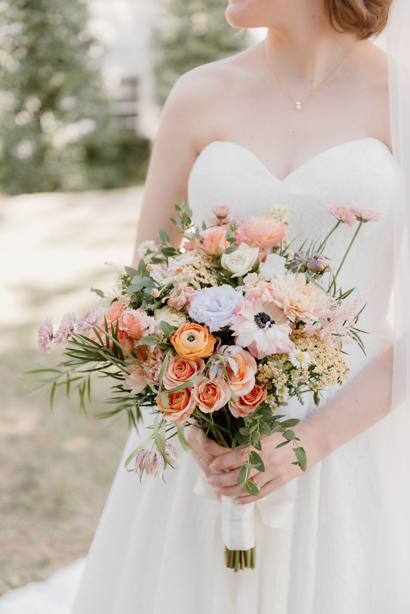 Bride in white strapless gown holding lush bridal bouquet with peach roses, ranunculus, anemones, and pink blooms.