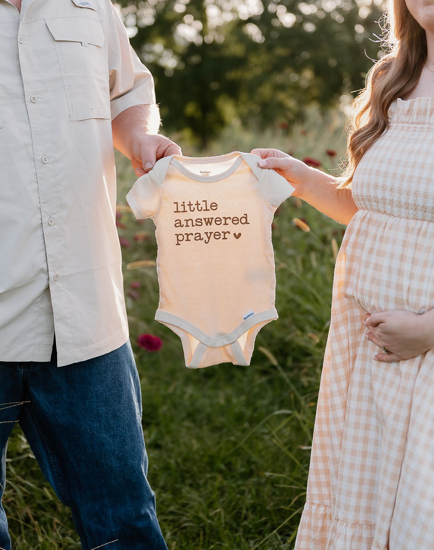 Couple holding baby onesies in a sunlit garden with gingham dress and white shirt.