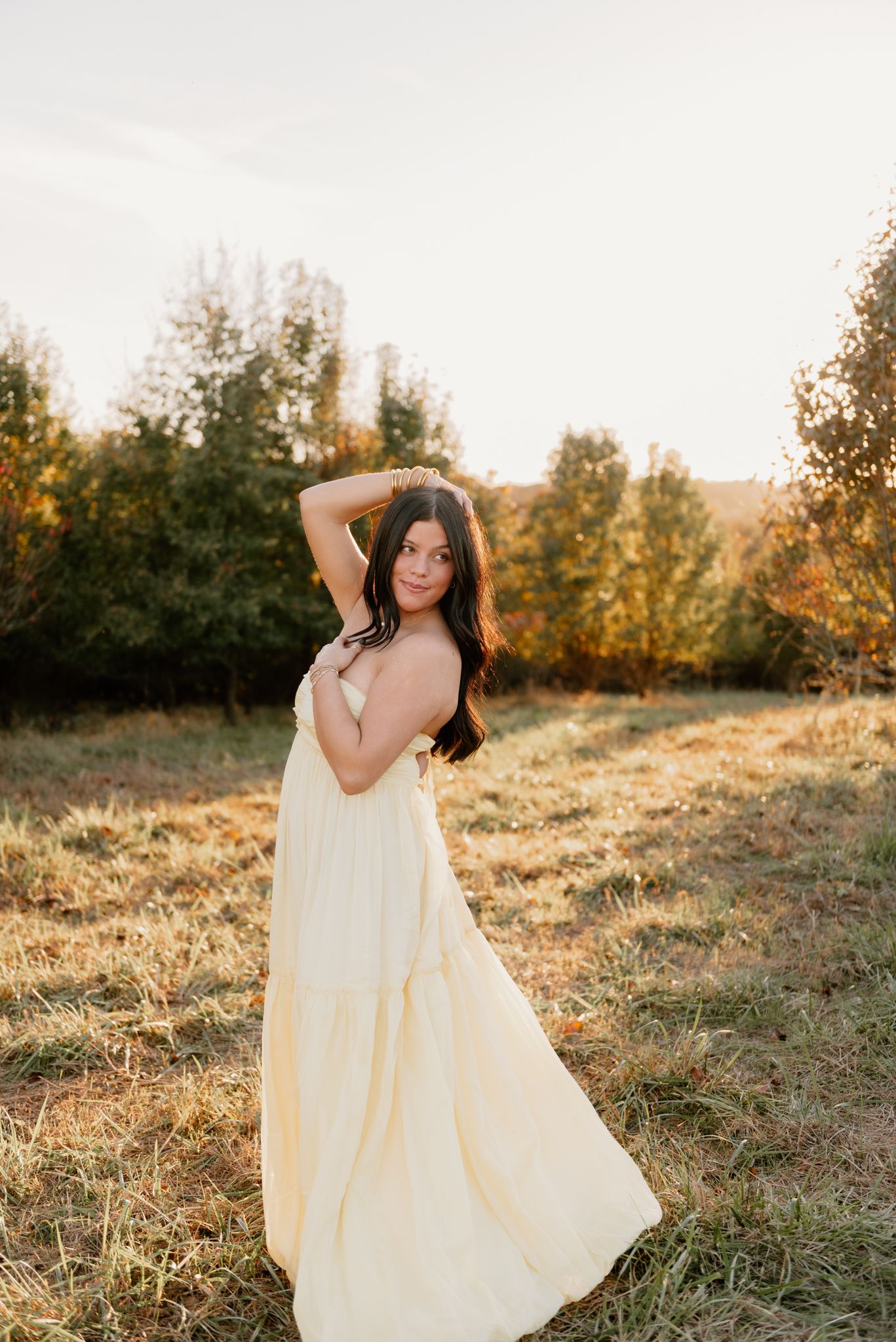 Young woman in flowing yellow dress poses gracefully in golden hour sunlight with autumn trees in background.