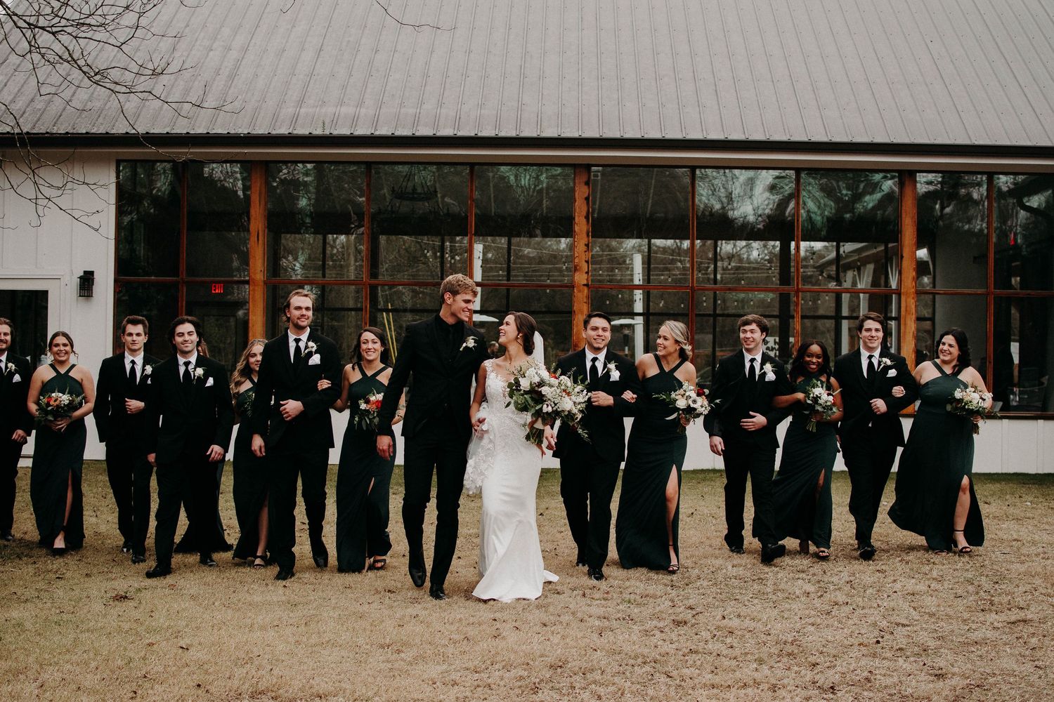 Wedding party in formal black and white attire lined up against rustic venue exterior.