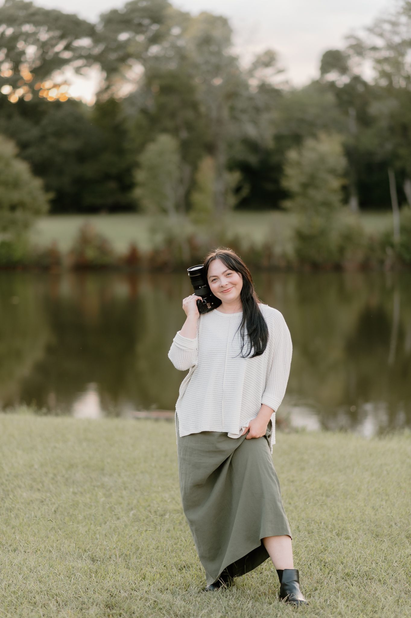 Woman in white top and olive skirt poses gracefully by a serene pond.