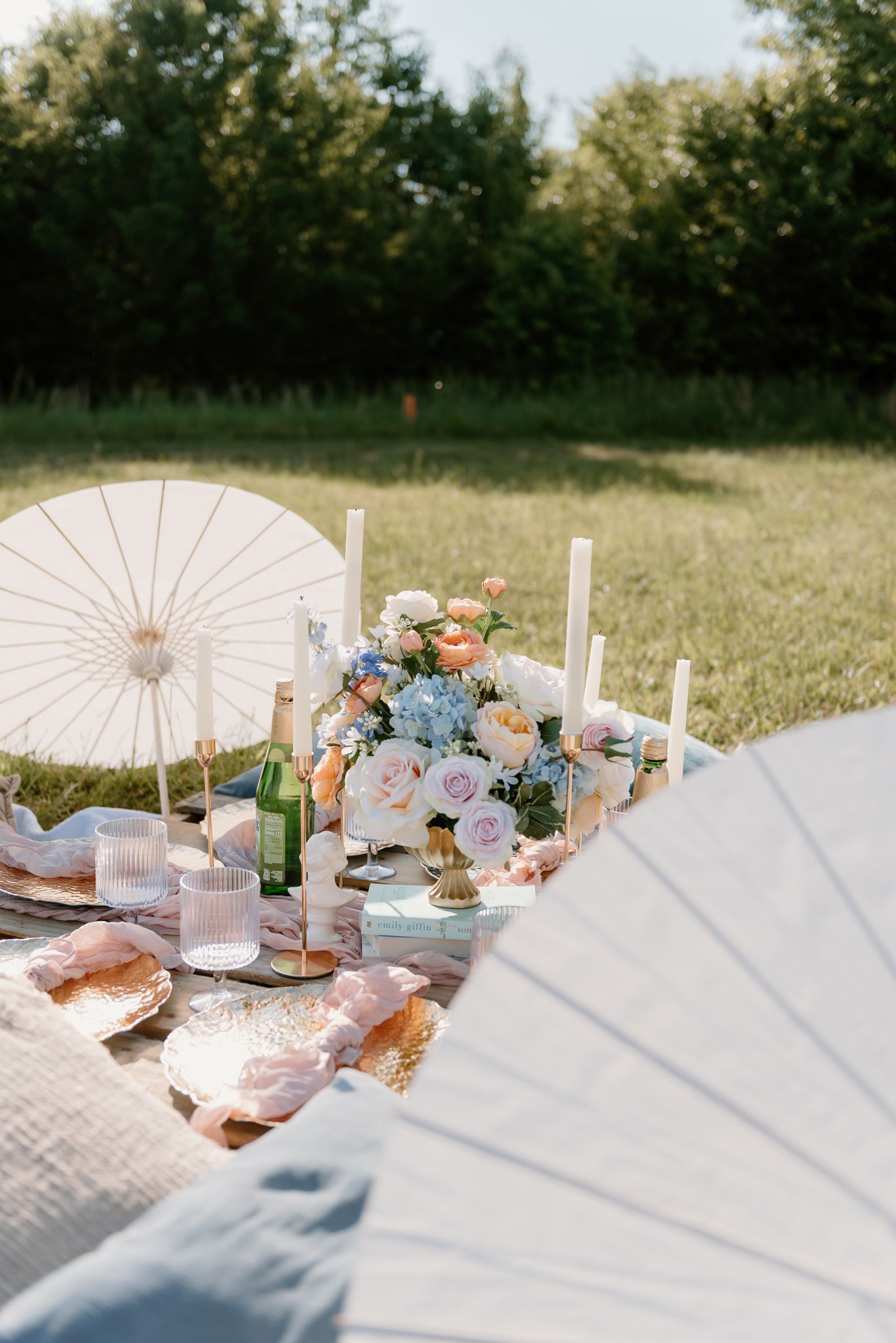 Elegant picnic setup with parasol, flowers, and candles on a blanket in a sunny field.