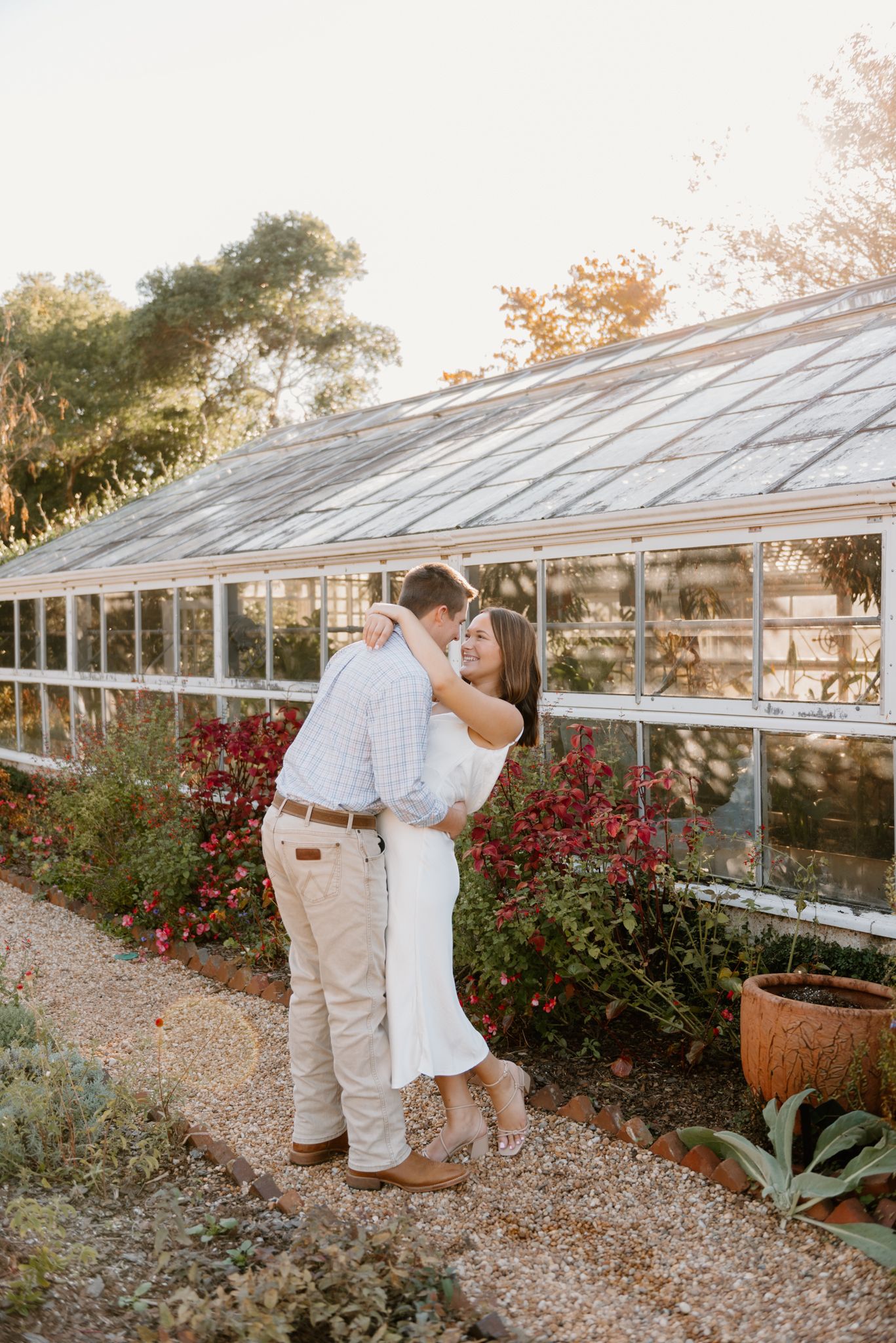 A romantic couple embraces near a rustic greenhouse surrounded by red flowers and gravel paths on a sunny afternoon.