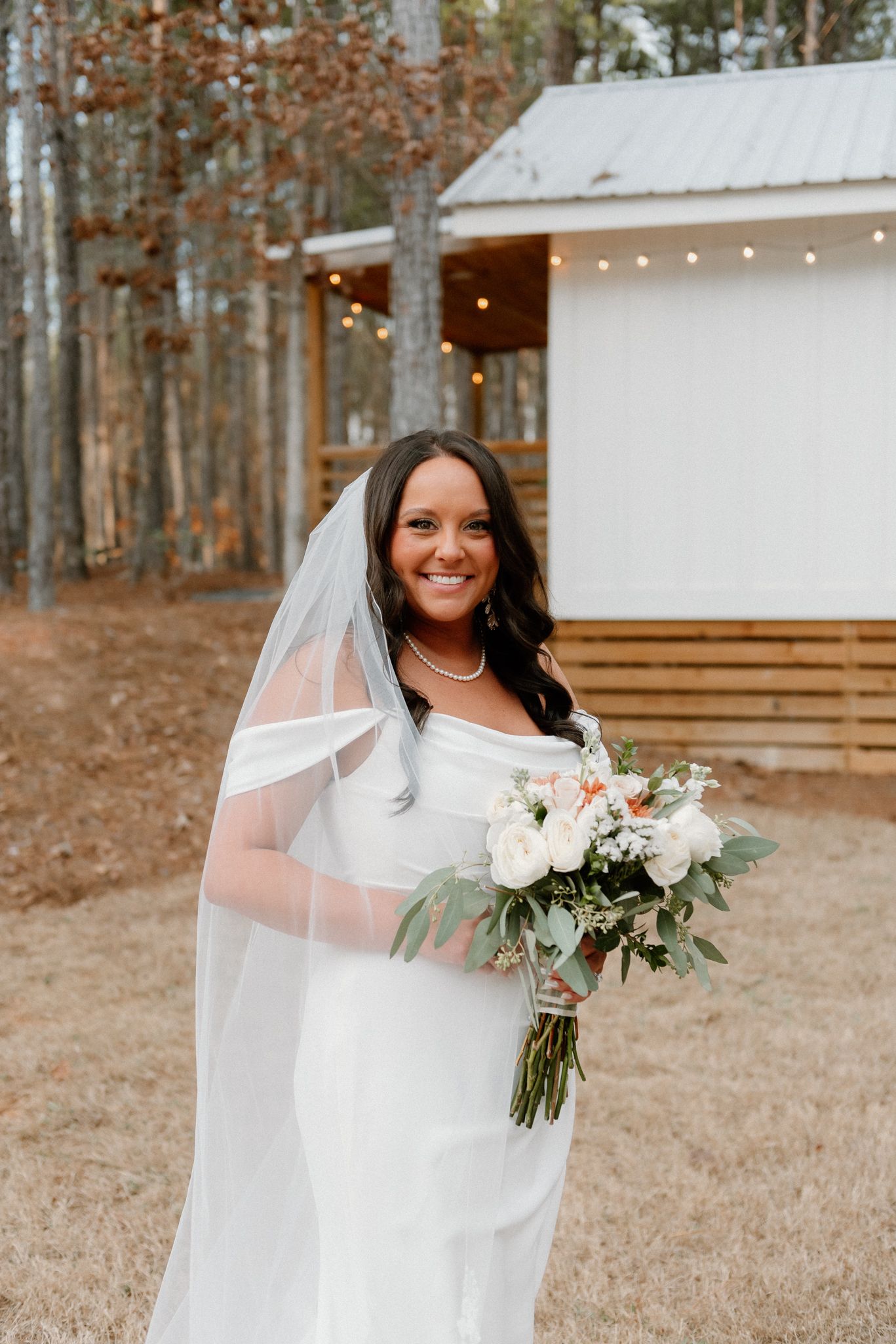 Bride in white dress and veil holding white floral bouquet outside rustic venue.