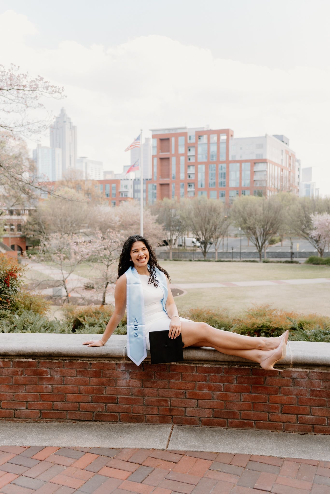 A smiling woman in graduation attire sits on a brick wall with cherry blossoms and city skyline behind her.