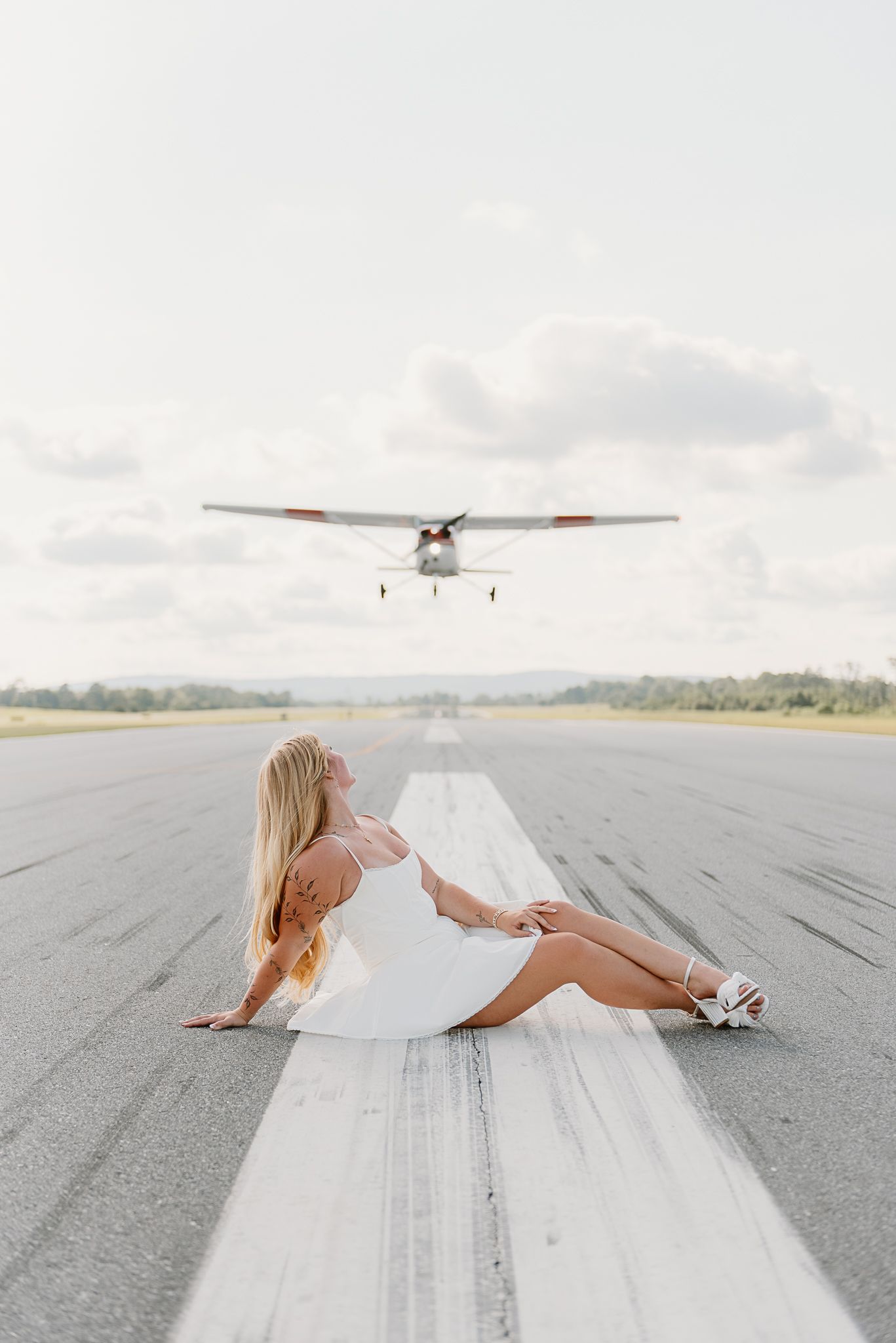 A person in white dress poses on an airport runway with a small aircraft in the background.