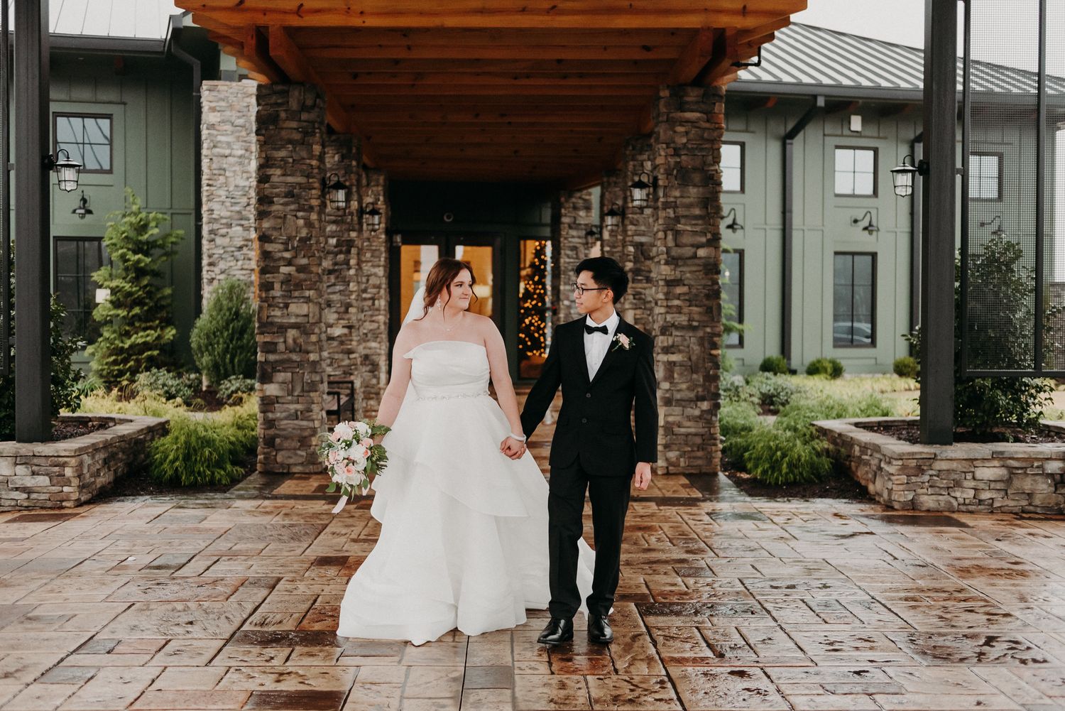Elegant couple walking along stone pathway outside rustic venue.
