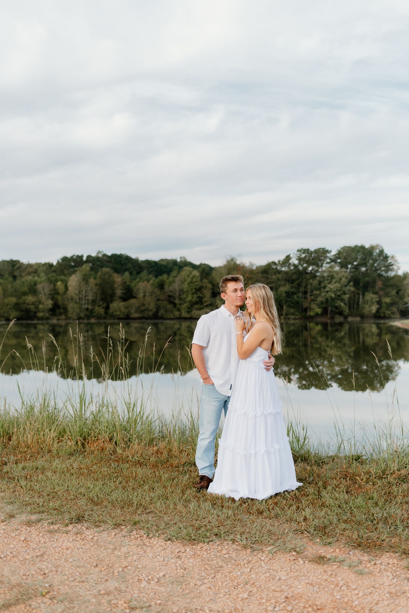 Couple embracing by a serene lake, woman in white dress and man in white shirt and jeans, lush trees reflecting on water.
