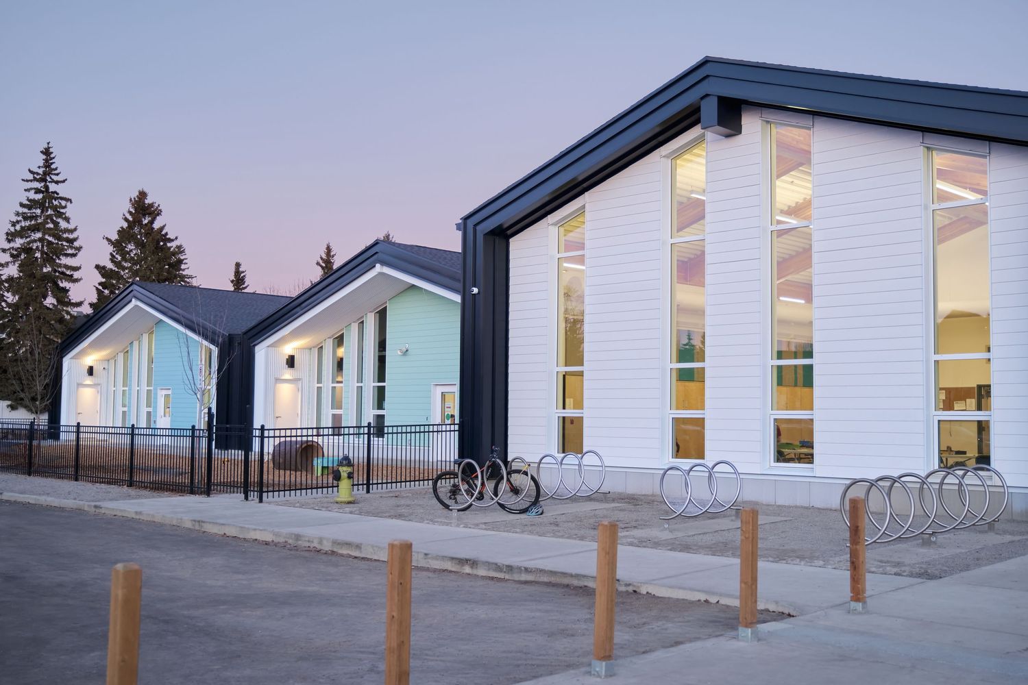Modern school building with white siding and blue accents, bike racks, and wooden bollards at dusk.