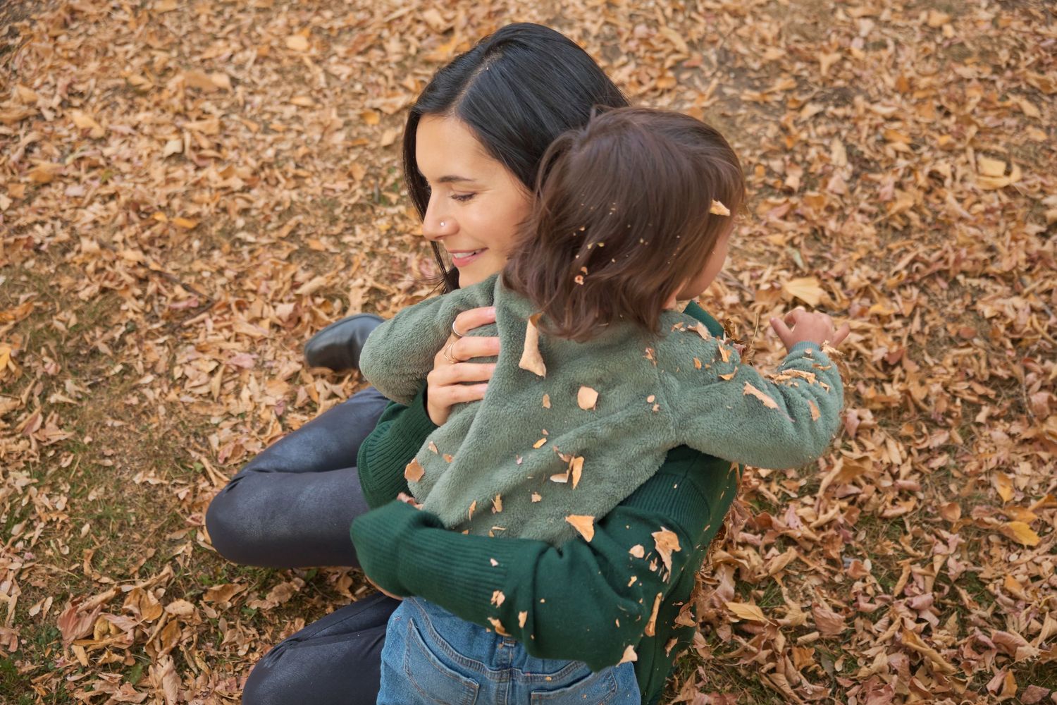 Two people embrace warmly while sitting on fallen autumn leaves in a park setting.