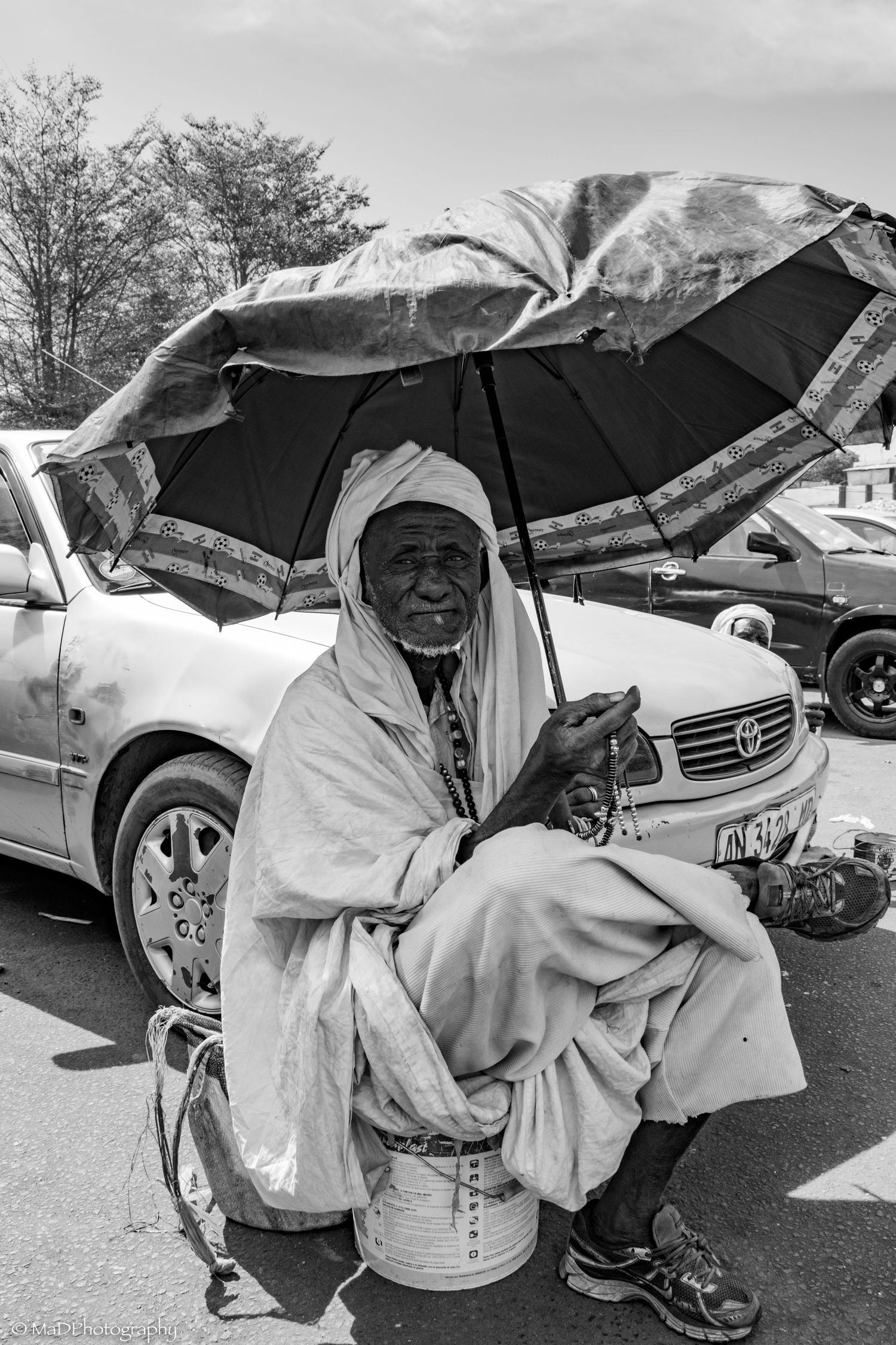 Black and white portrait of person with umbrella sitting near parked cars.