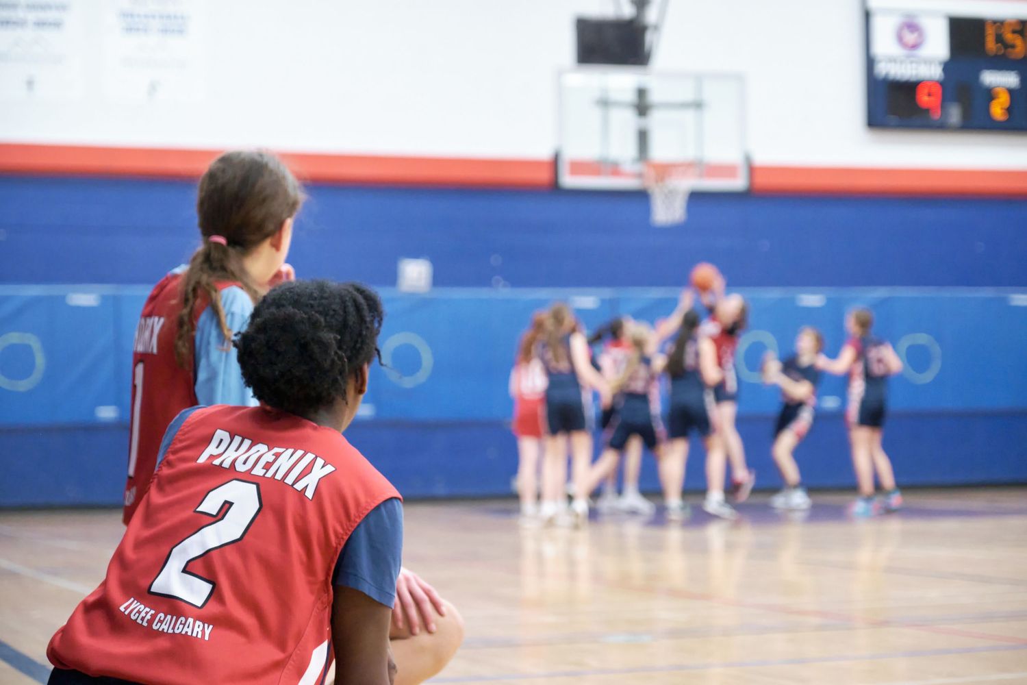 Two youth basketball players watch teammates celebrate on an indoor court, player wearing Phoenix number 2 jersey.