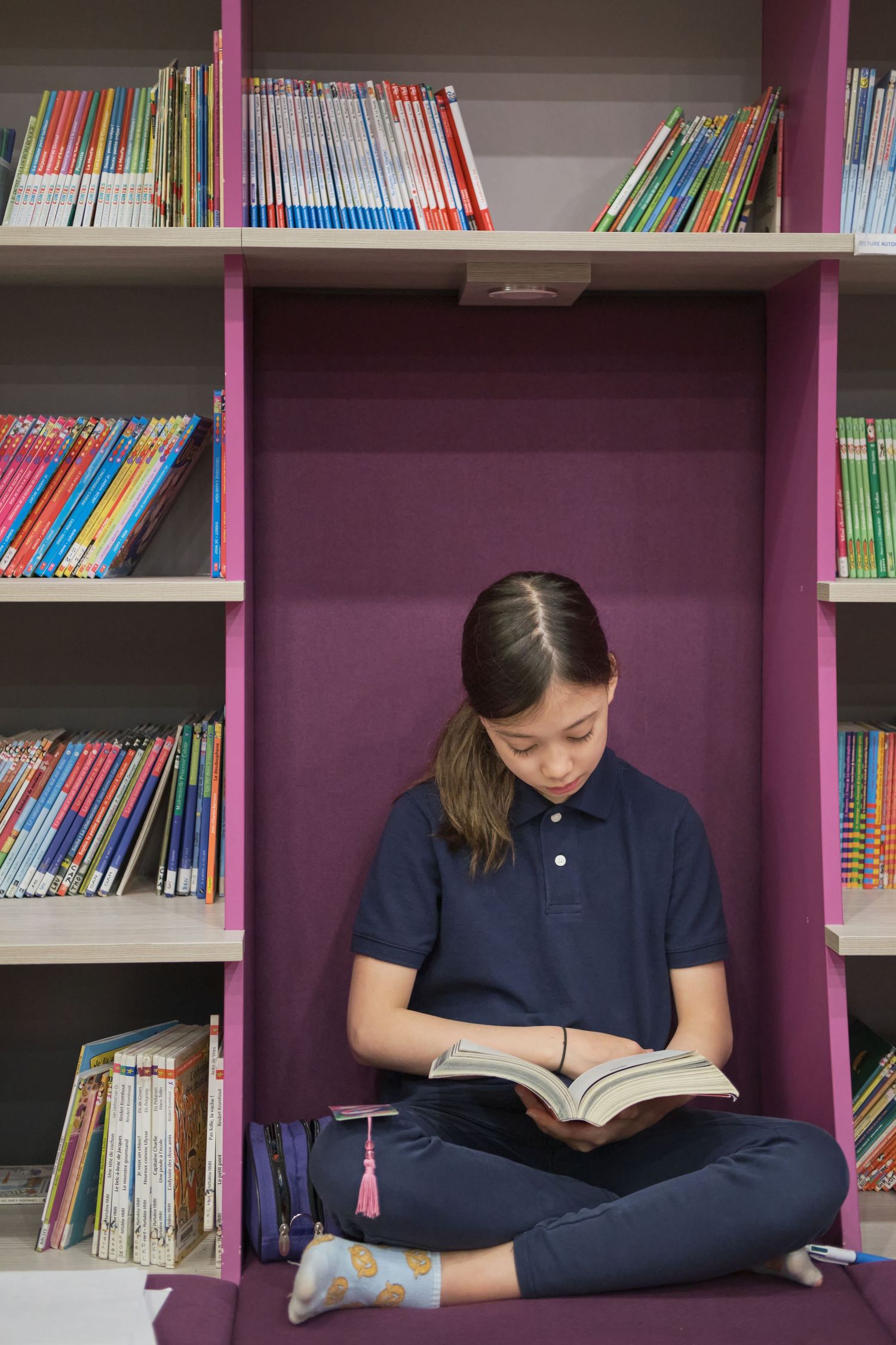 Student in navy blue uniform sits cross-legged on library shelf reading a book among colorful spines.