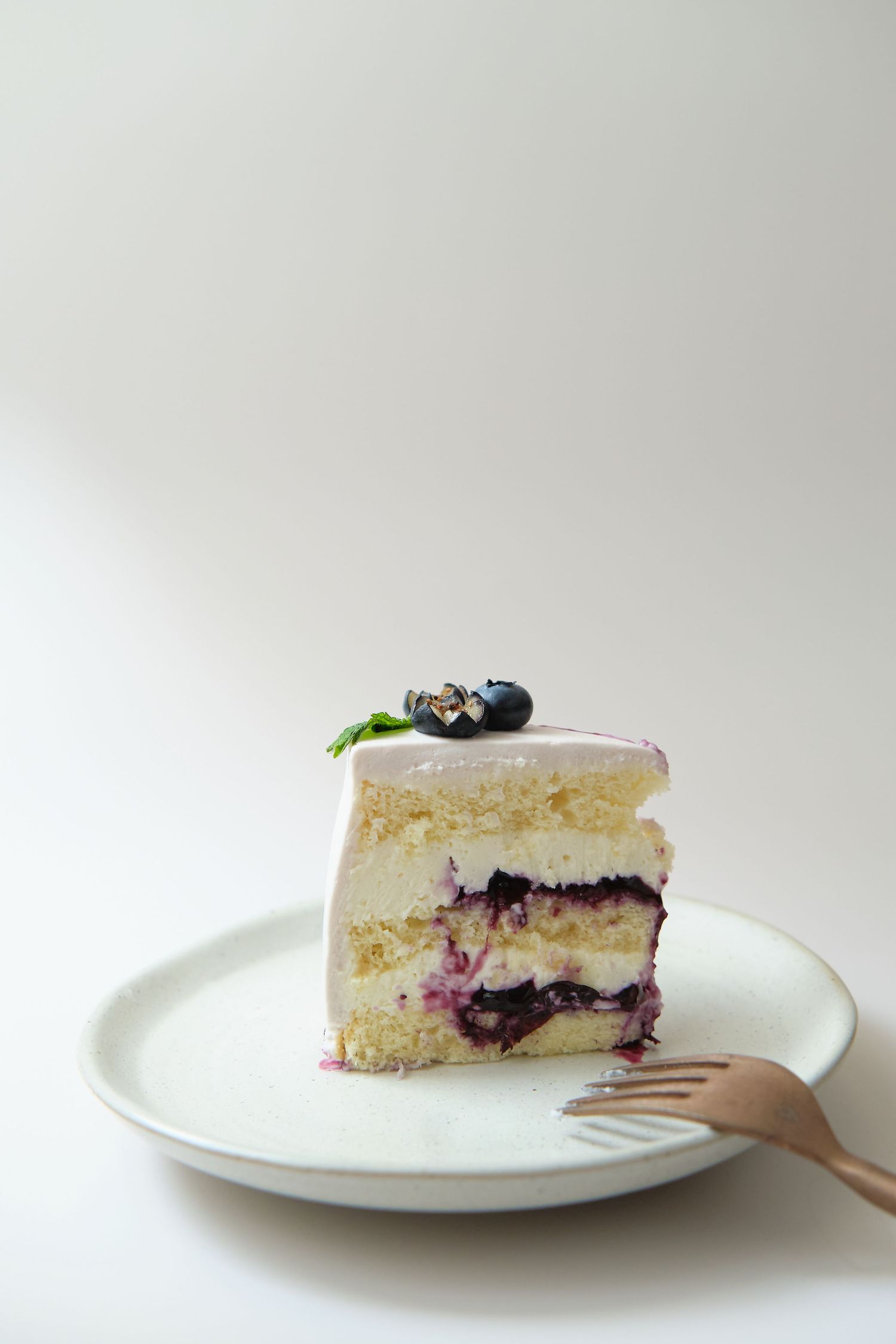 Slice of layered vanilla cream cheese cake with blueberry filling served on white plate.
