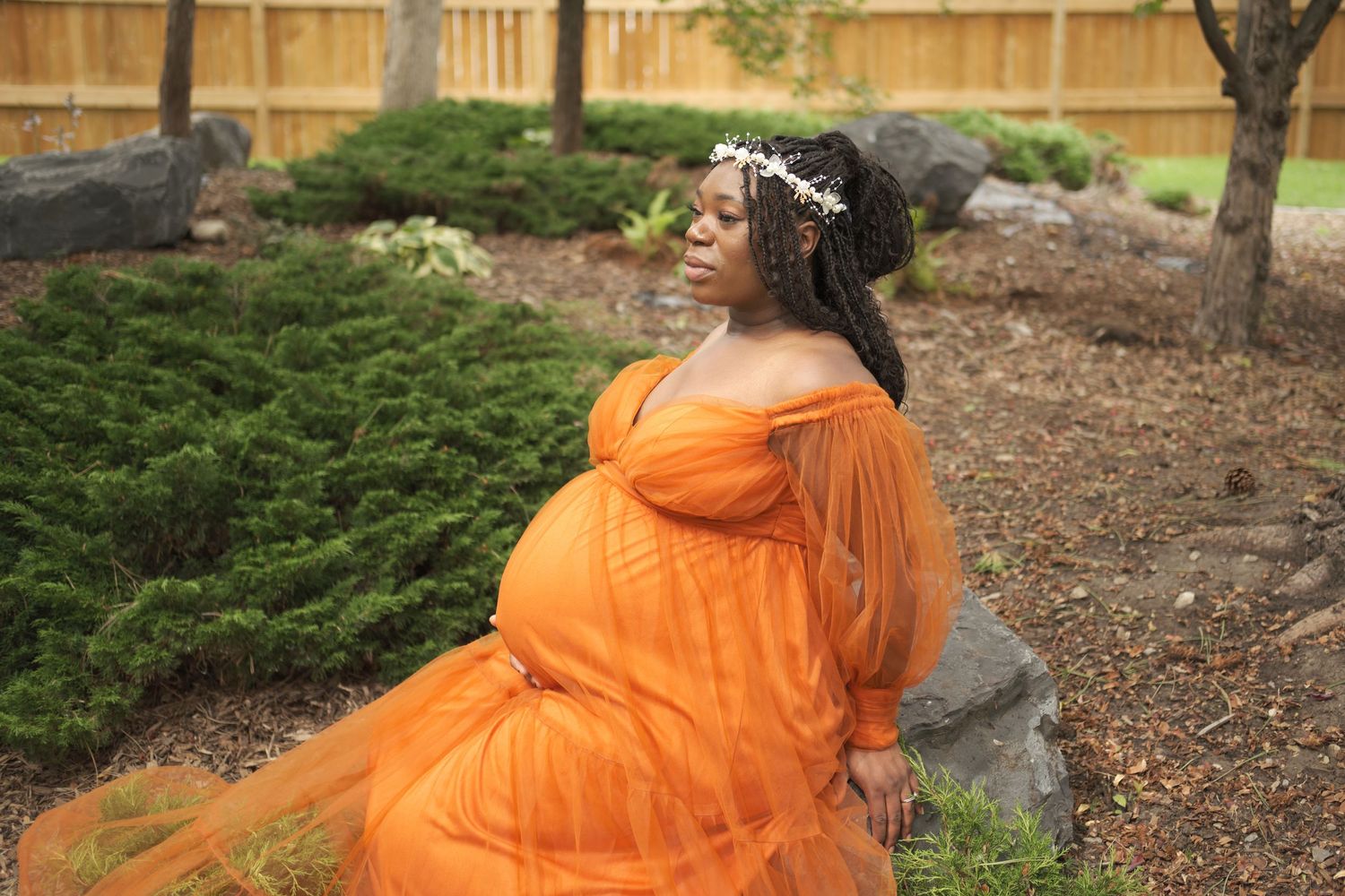Expecting mother in flowing orange dress and floral crown sits on rock in garden setting during maternity photoshoot.