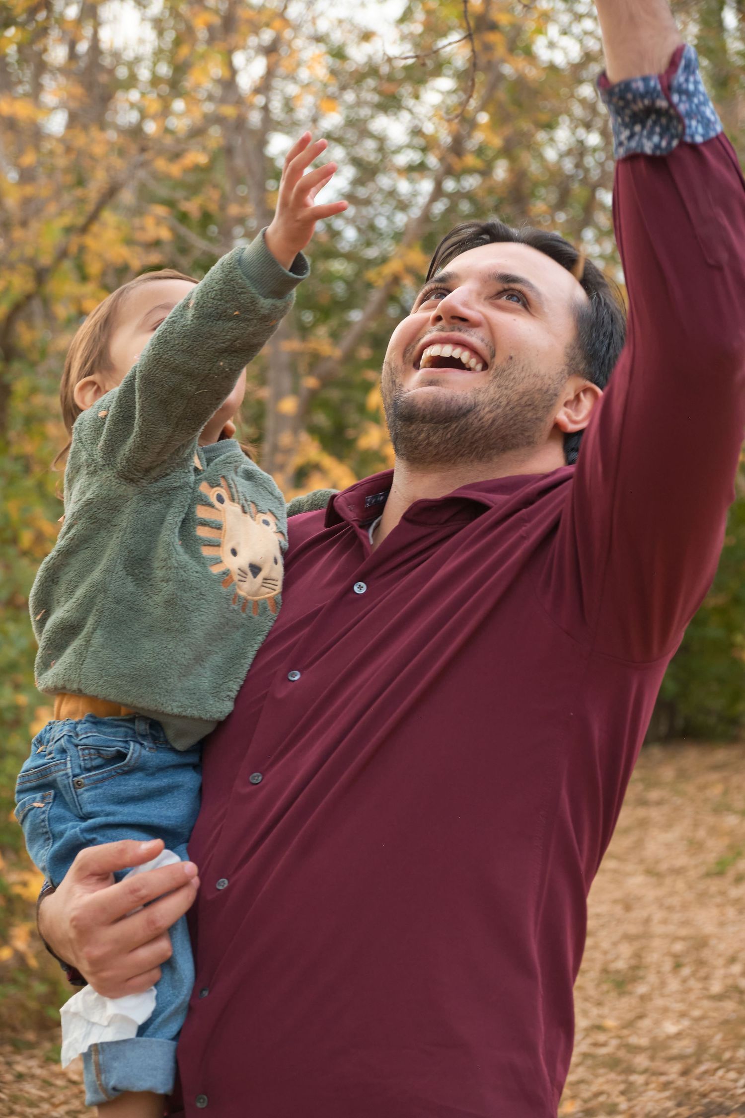 A parent in a burgundy shirt lifts and plays with a small child wearing green outdoors during autumn.