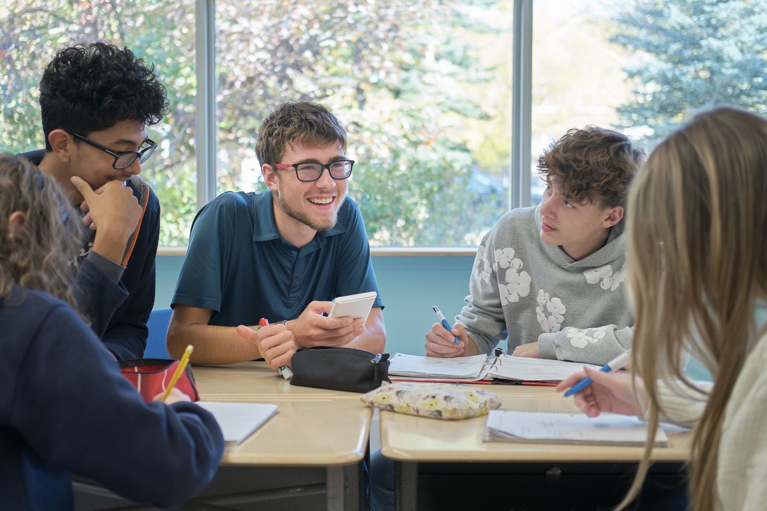Students work together at desks in a bright classroom setting with large windows.