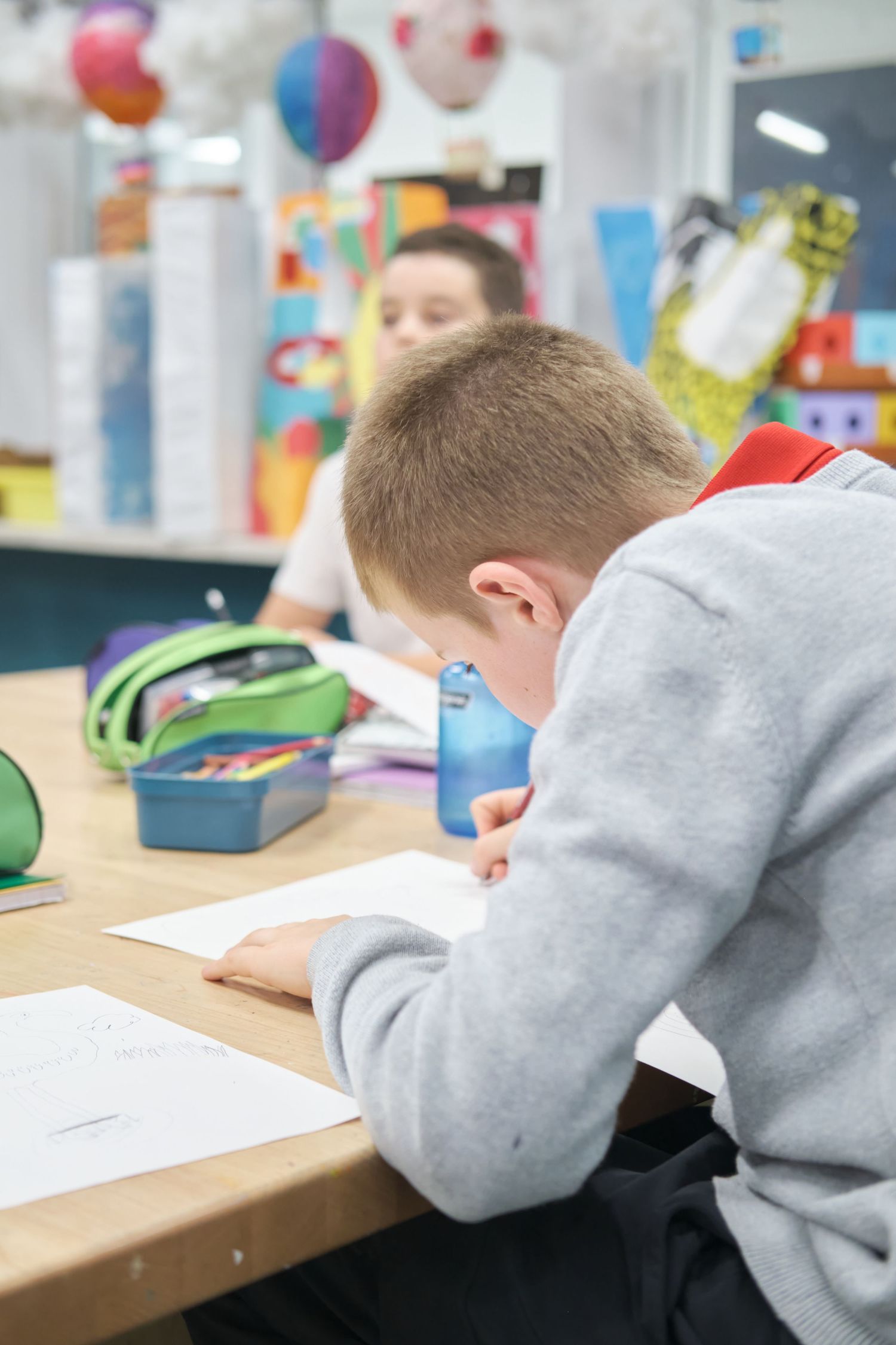 Students in gray sweatshirts sit at wooden desks working on papers in a classroom decorated with colorful balloons.