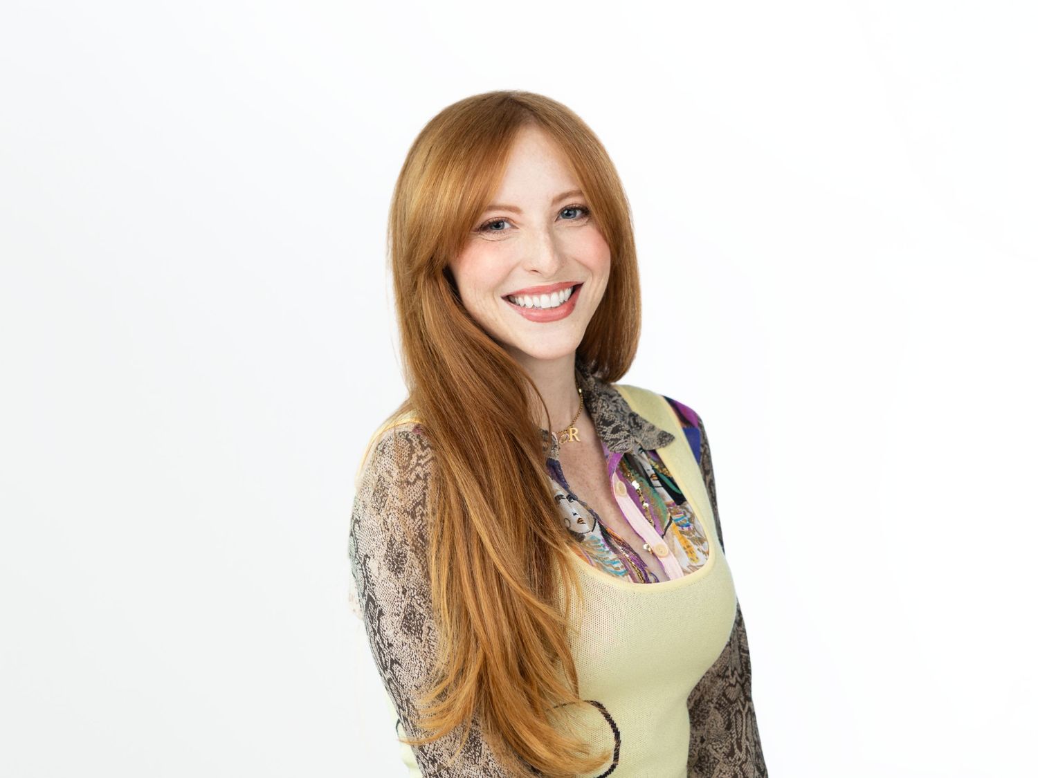 Smiling woman with long red hair wearing a light green top layered over a patterned snakeskin-print blouse.