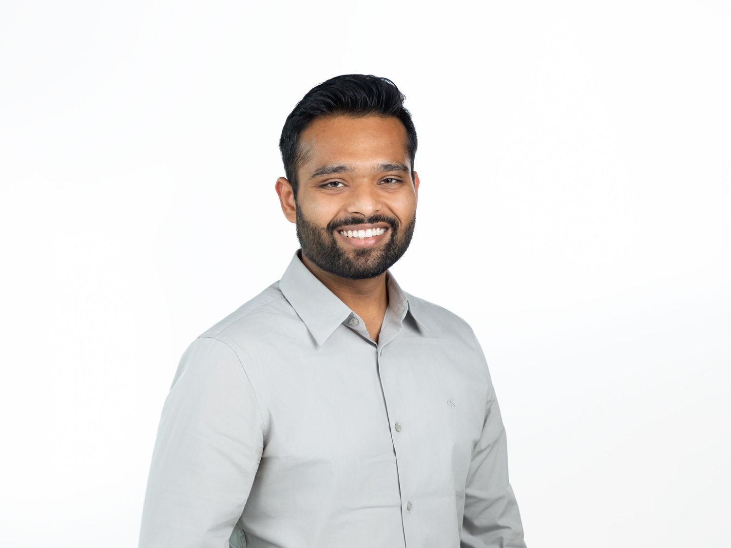 Smiling bearded man with short dark hair wearing a light gray button-up shirt against a clean white background.