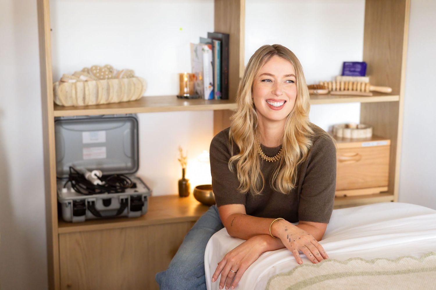 Smiling woman with long blonde hair sits on a treatment table in a cozy wellness room with wooden shelves and a portable device.