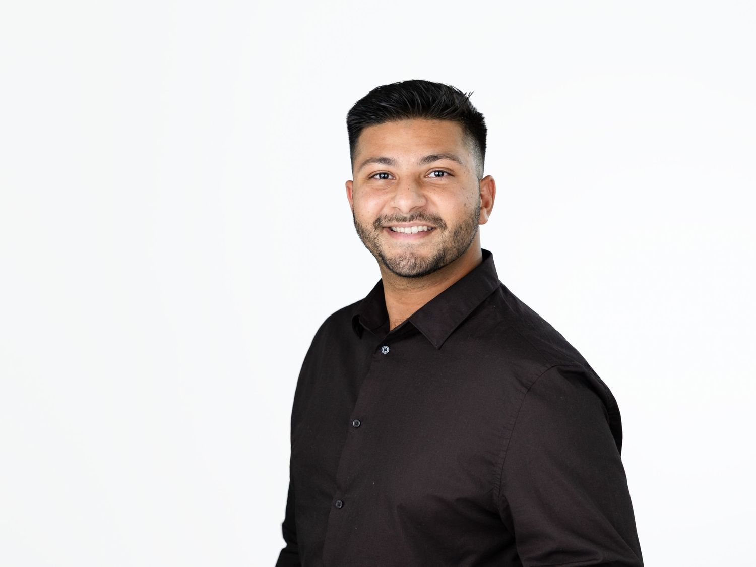 Smiling man with short dark hair and a trimmed beard wearing a black button-up shirt against a white studio background.