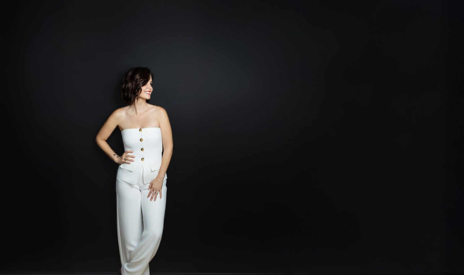 Woman with short dark hair stands with a hand on her hip in a white strapless jumpsuit against a black studio backdrop.