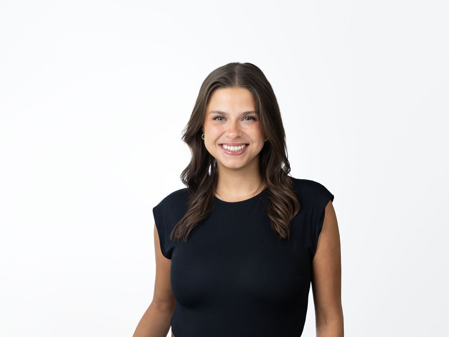 Smiling woman with long wavy brown hair wearing a black sleeveless top posing against a clean white background.