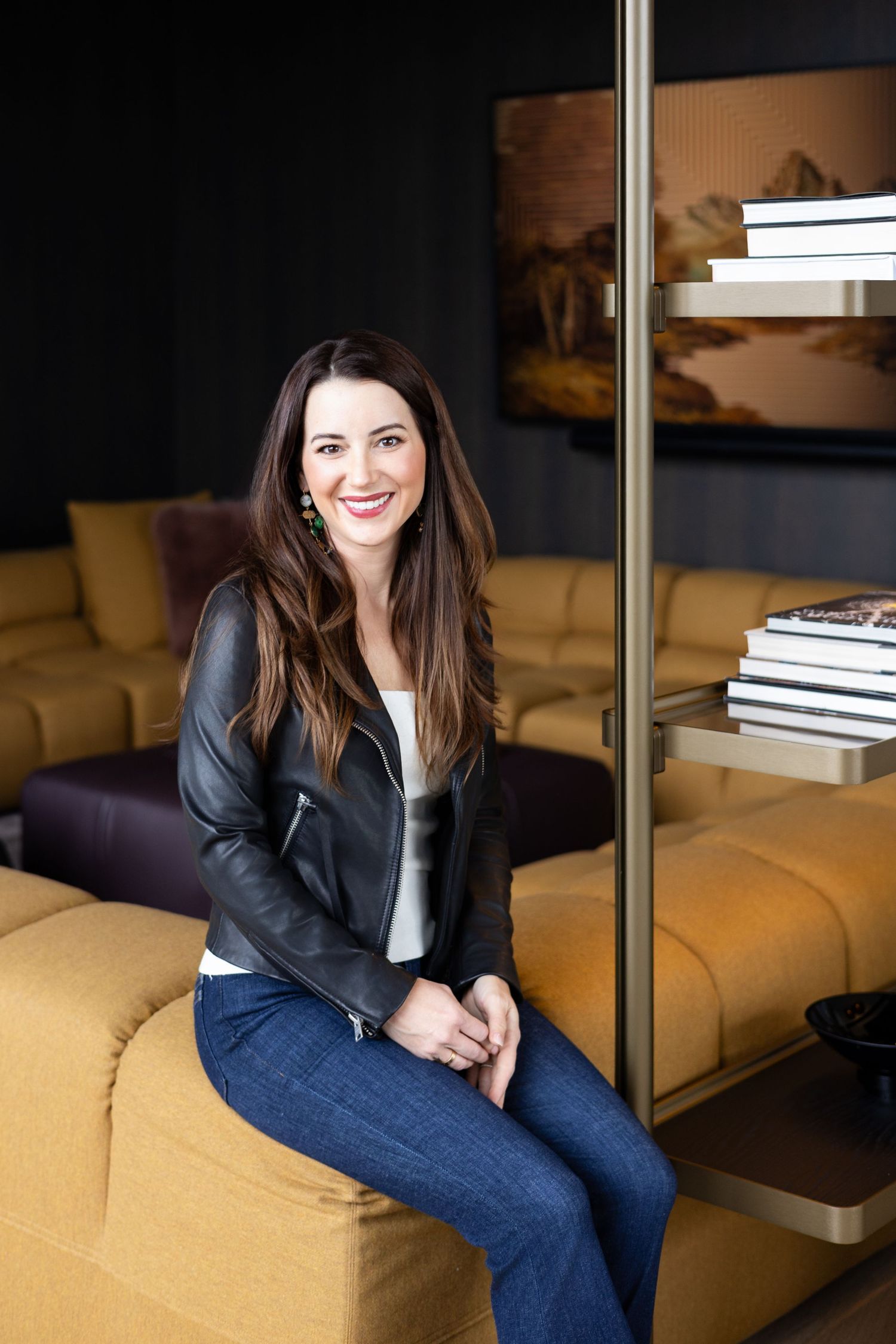 A smiling woman in a black leather jacket and jeans sits on a mustard yellow sofa in a modern, dark-toned lounge.