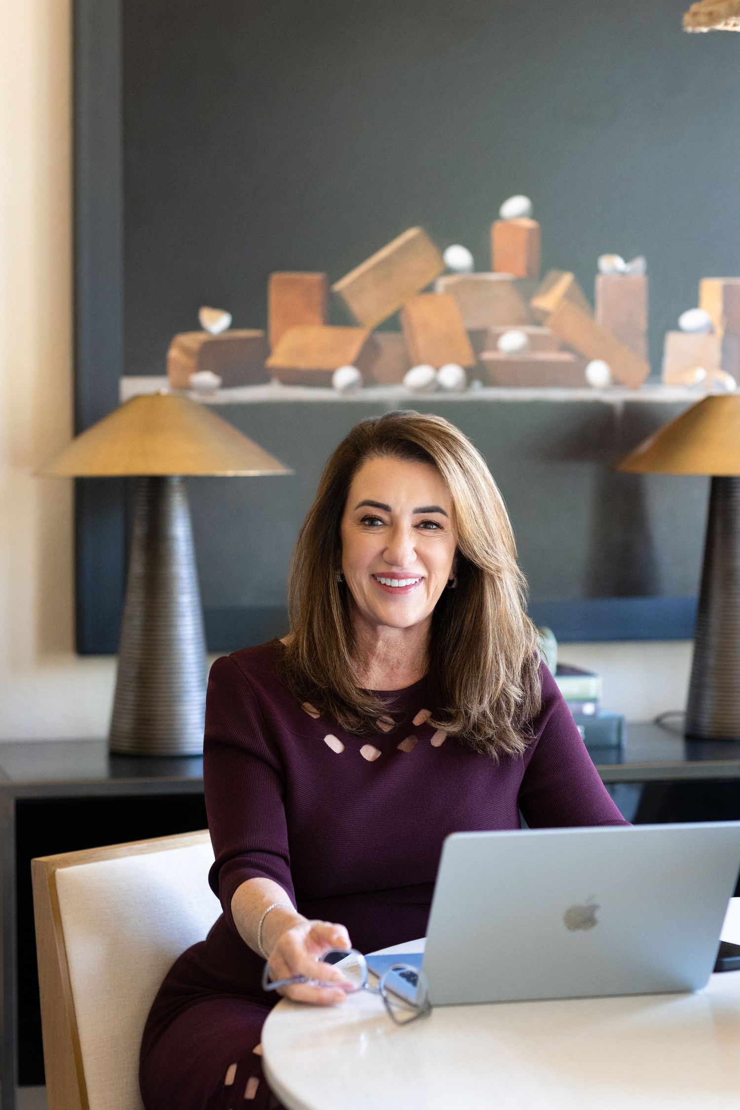 Smiling woman in a burgundy top sits at a white table with an open laptop and glasses in a stylish office with sculptural art behind her.