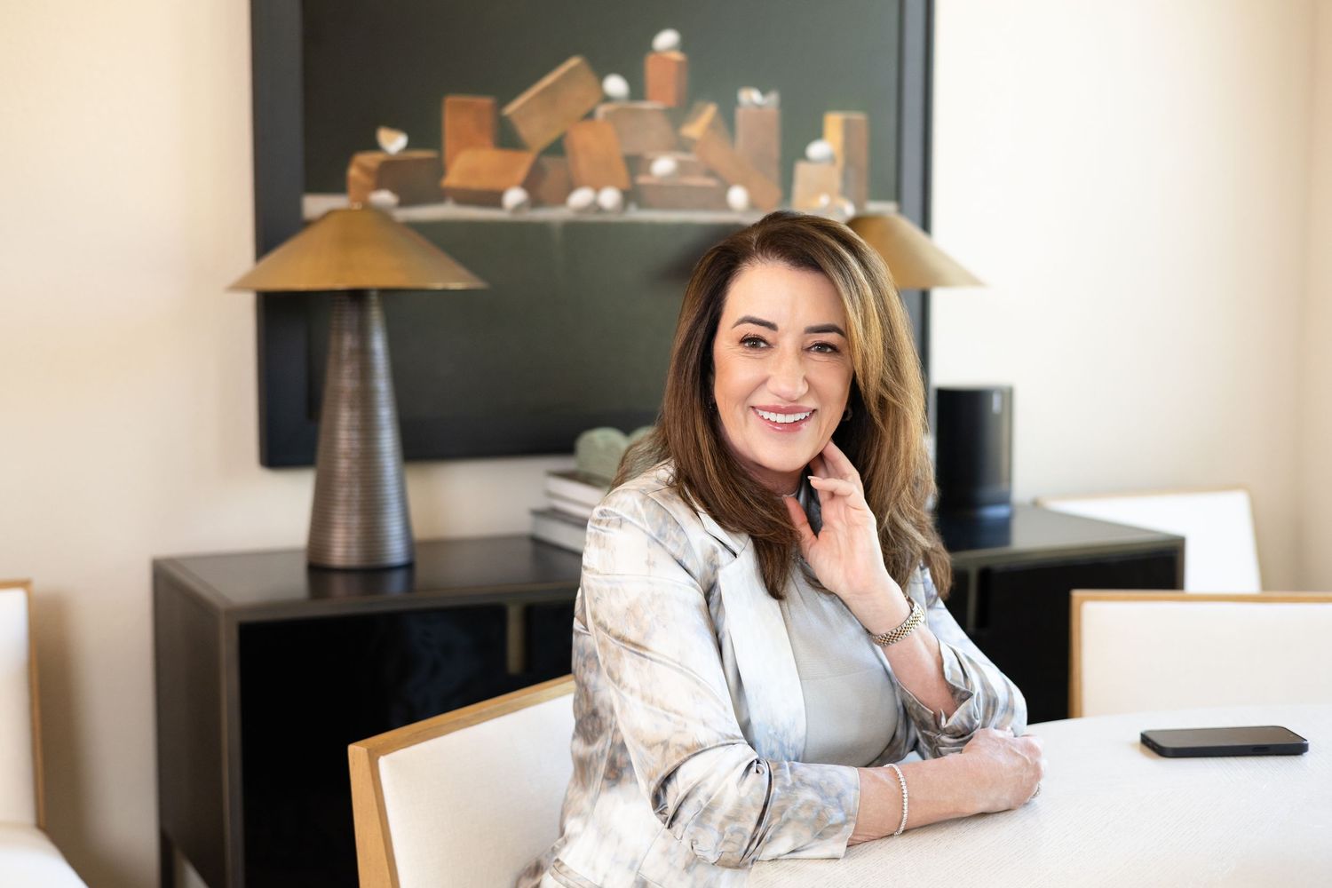 Woman in a light patterned blazer rests her hand near her face and smiles while seated at a white table in an elegant modern office.