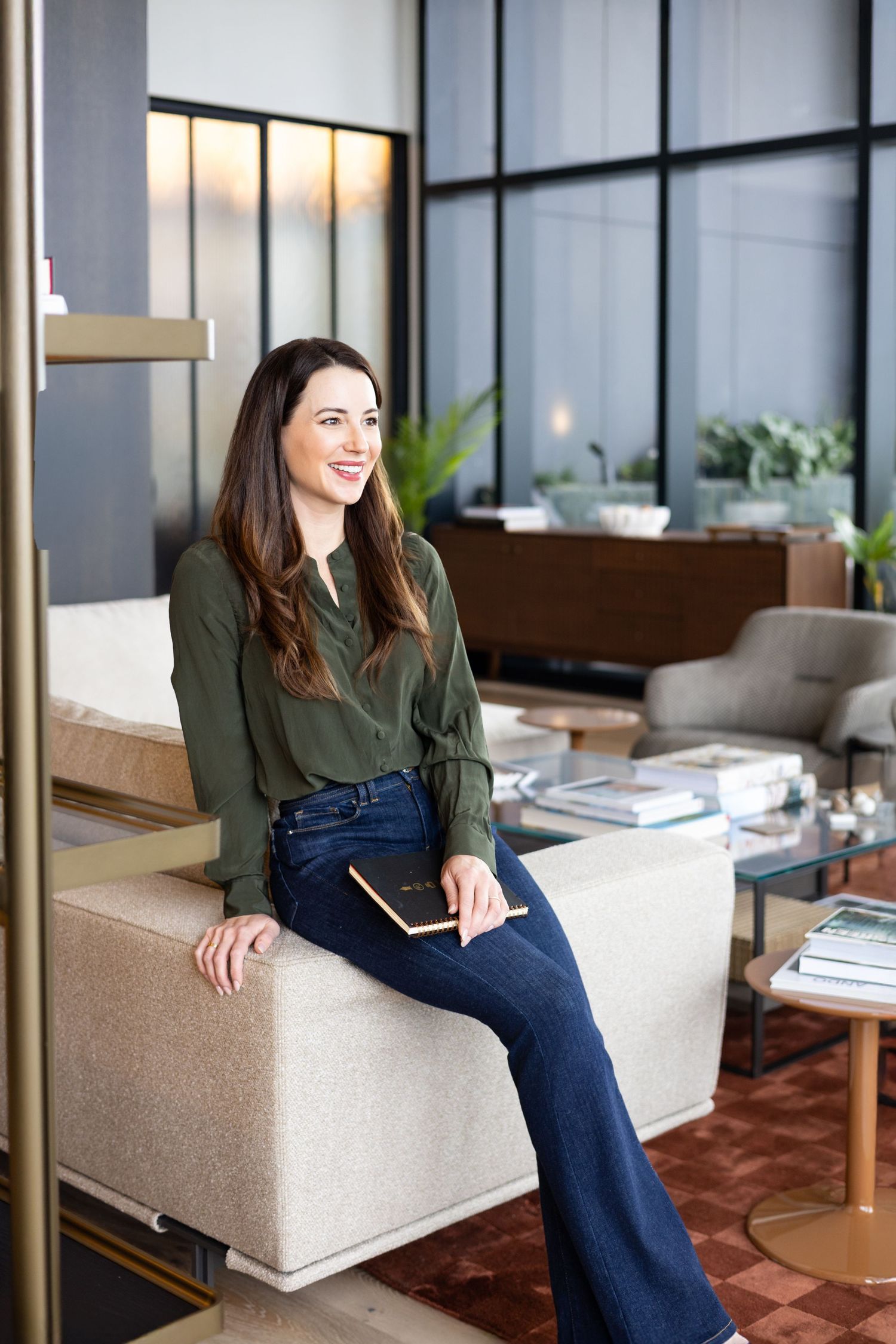 A smiling woman in a green blouse and flared jeans sits on a cream sofa holding a notebook in a modern, airy lounge.
