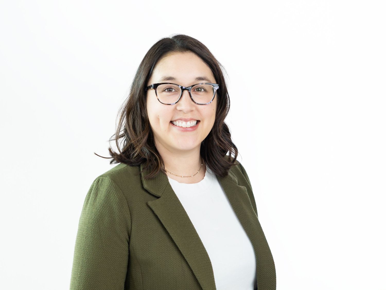 Smiling woman with dark wavy hair and glasses wearing an olive green blazer over a white top against a white background.
