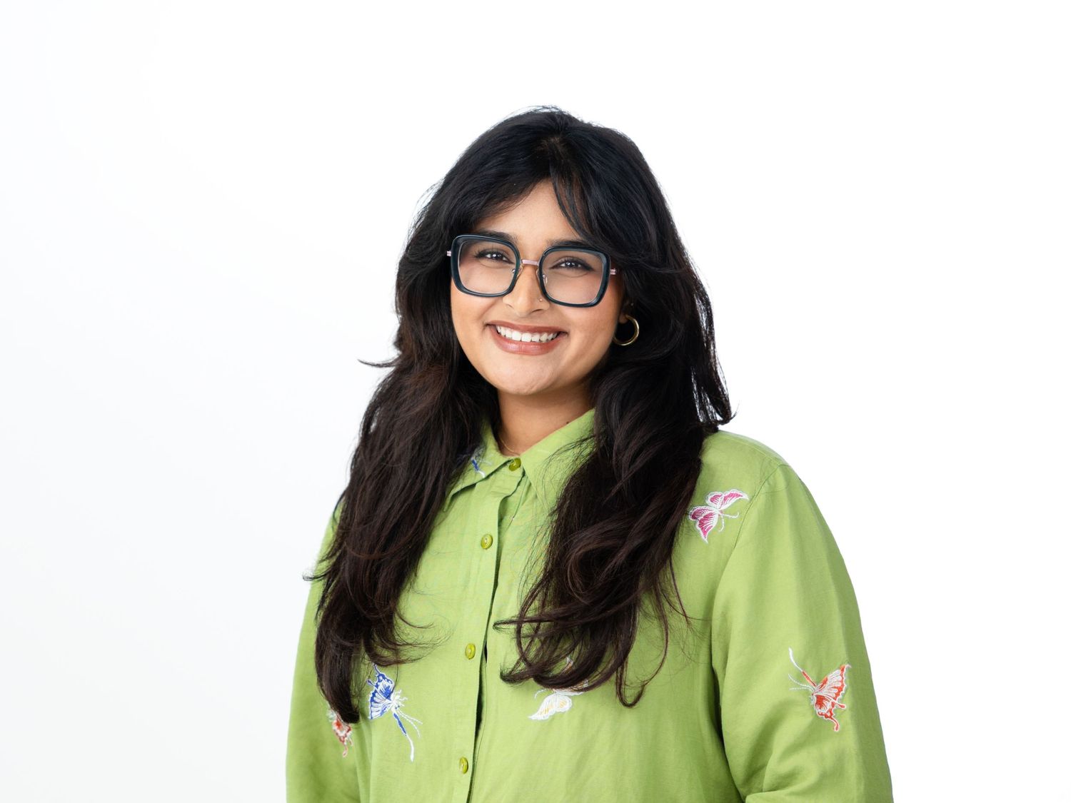 Smiling woman with long dark hair and glasses wearing a lime green embroidered button-up shirt against a white background.