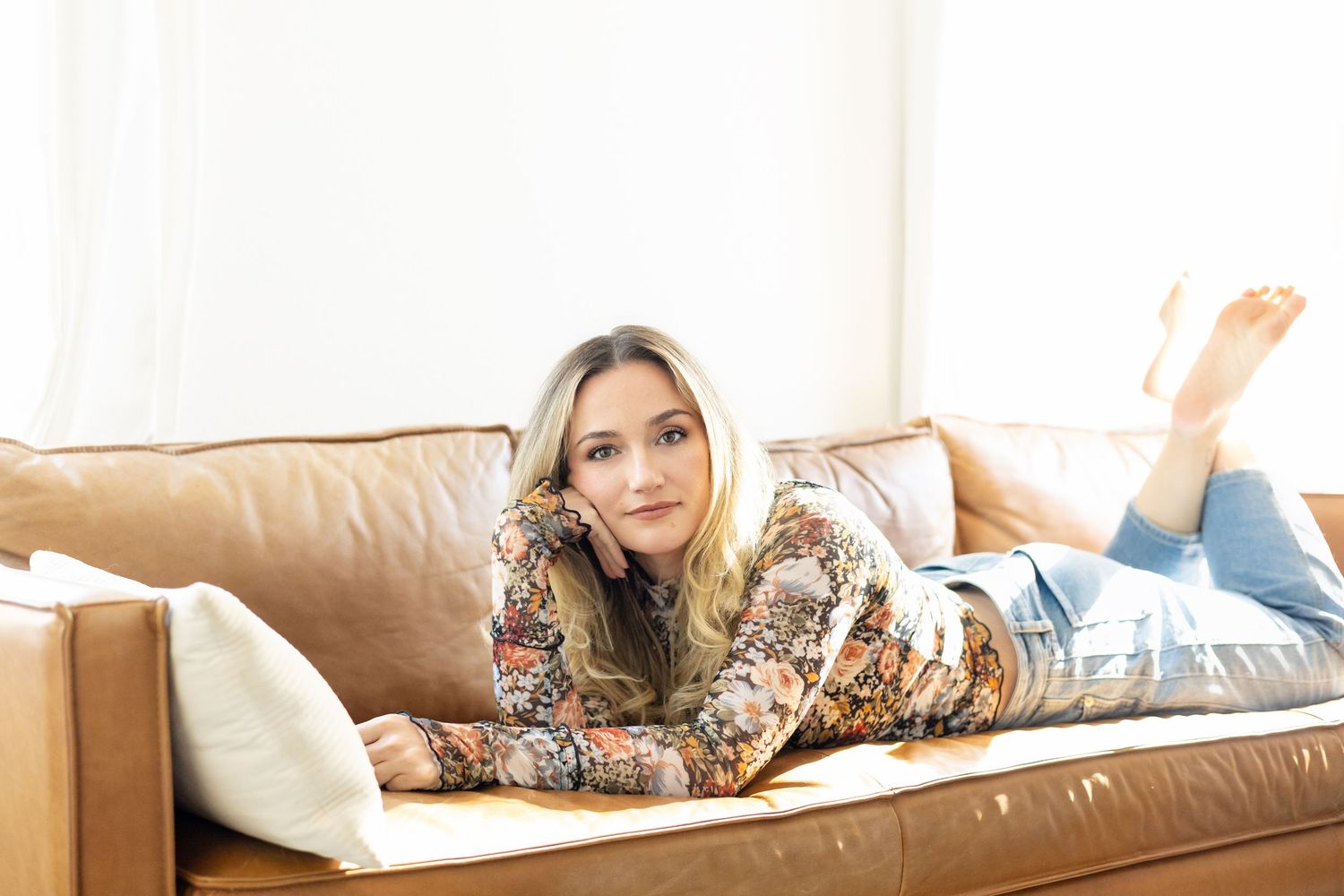 A woman with long blonde hair relaxes on a tan leather sofa in a bright room, wearing a floral top and jeans.