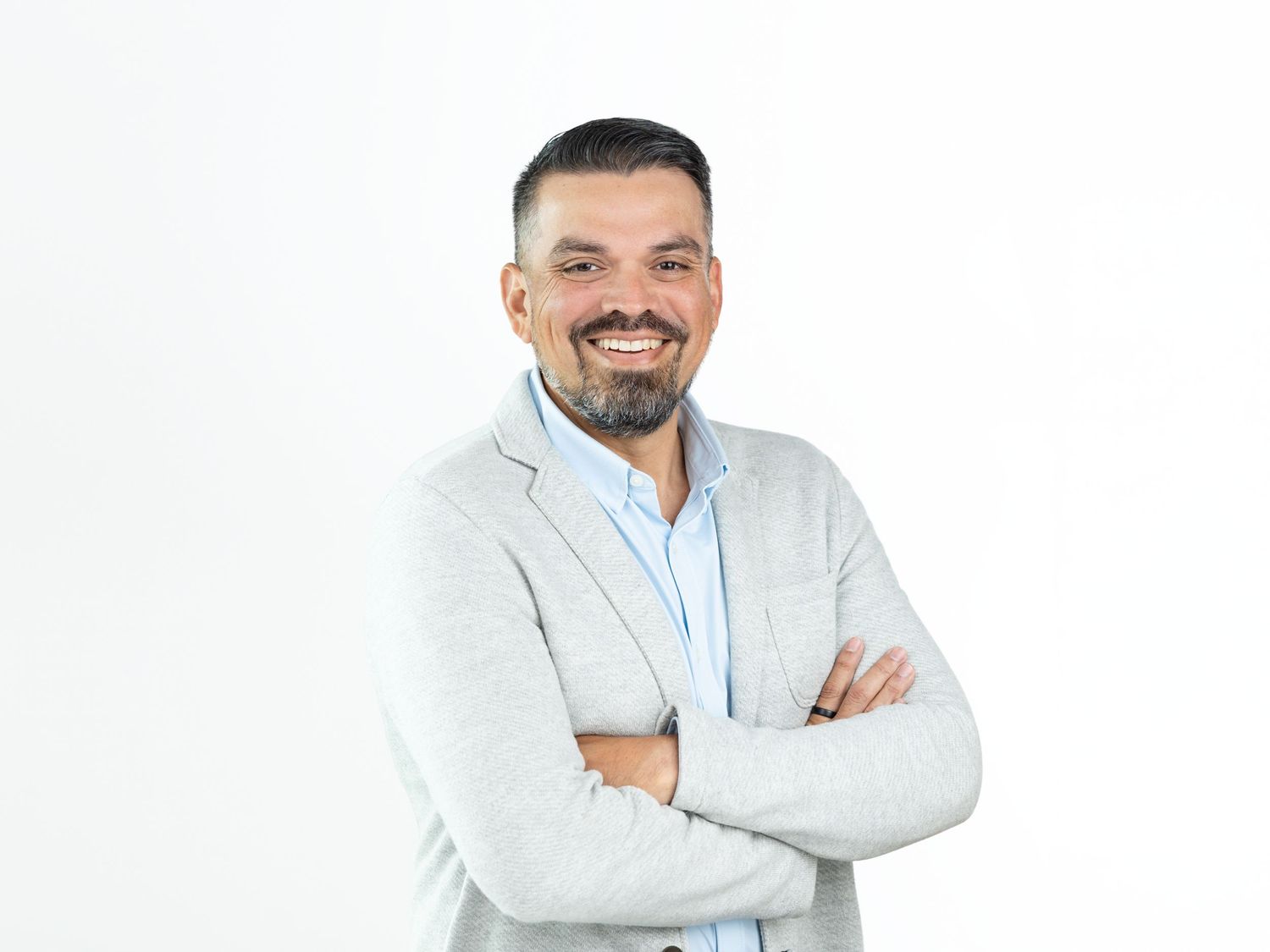 Smiling bearded man with crossed arms wearing a light gray blazer over a light blue collared shirt.