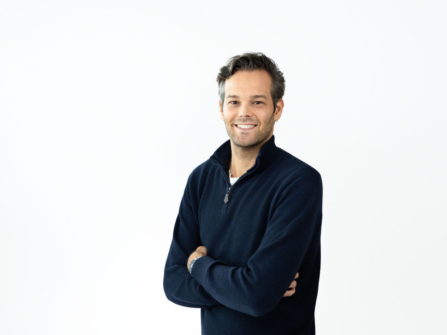 Smiling man with short dark hair and crossed arms wearing a navy quarter-zip sweater in a bright studio setting.