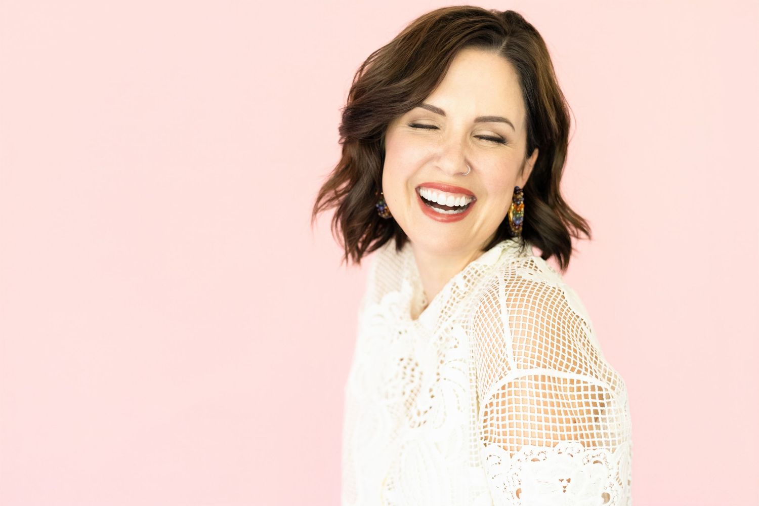 Woman with short wavy brown hair laughs with her eyes closed, wearing a white lace blouse and drop earrings on a pink background.