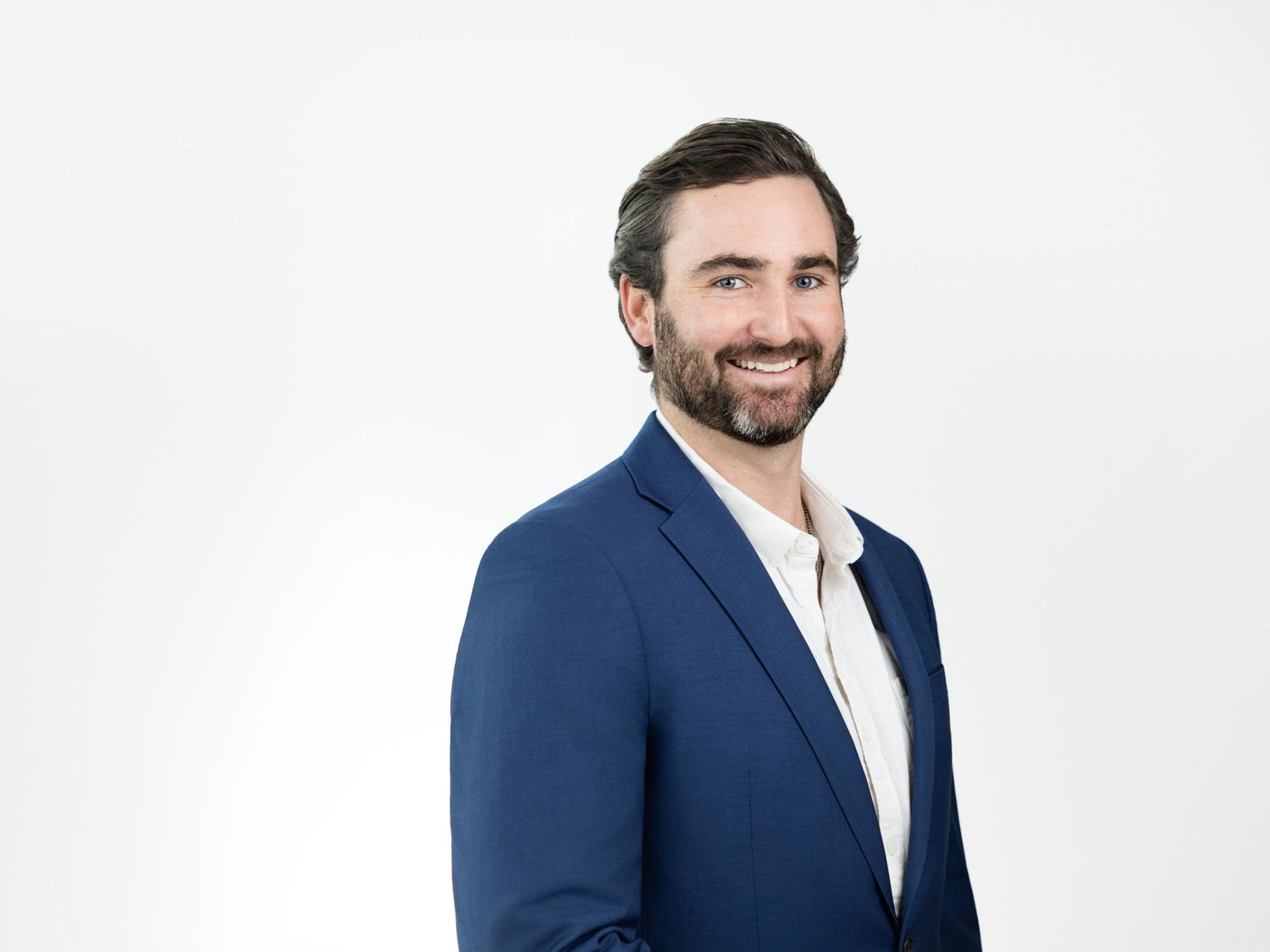 Smiling bearded man with short brown hair wearing a blue blazer over a white shirt posing against a white background.