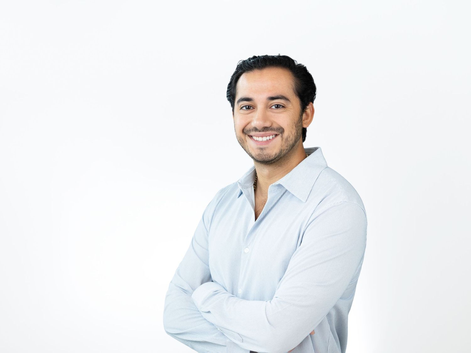 Smiling man with short dark hair and stubble stands with arms crossed in a light blue button-up shirt against a white background.