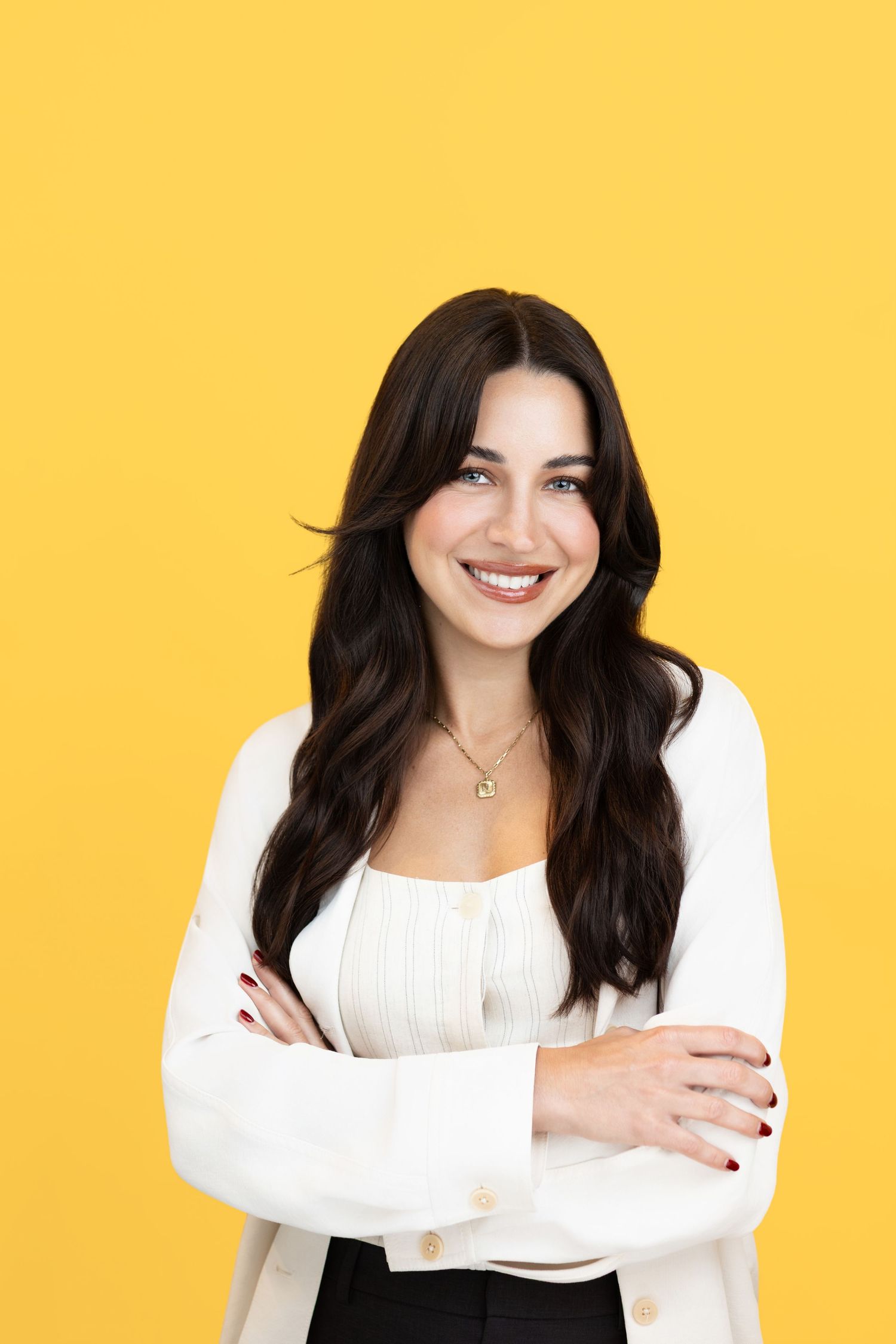 Smiling woman with long dark wavy hair and arms crossed wearing a white blazer against a bright yellow background.