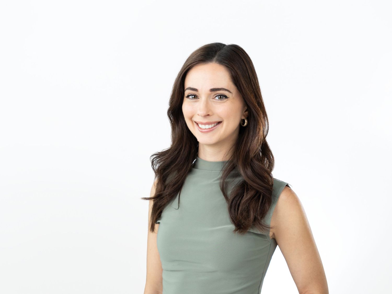 Smiling woman with long wavy brown hair and small earrings wears a sage green sleeveless top in a studio portrait on white.