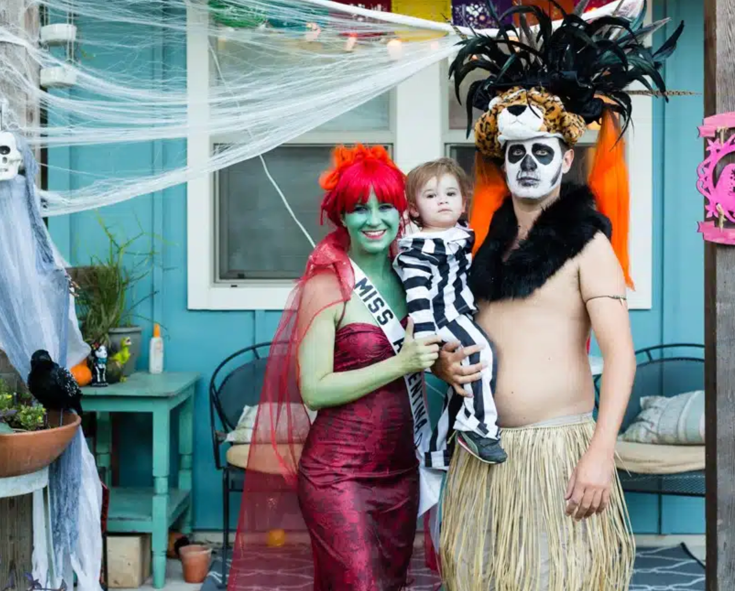 Family in Halloween costumes with skull face paint, a green-faced beauty queen and a toddler posing by a decorated porch with cobwebs.