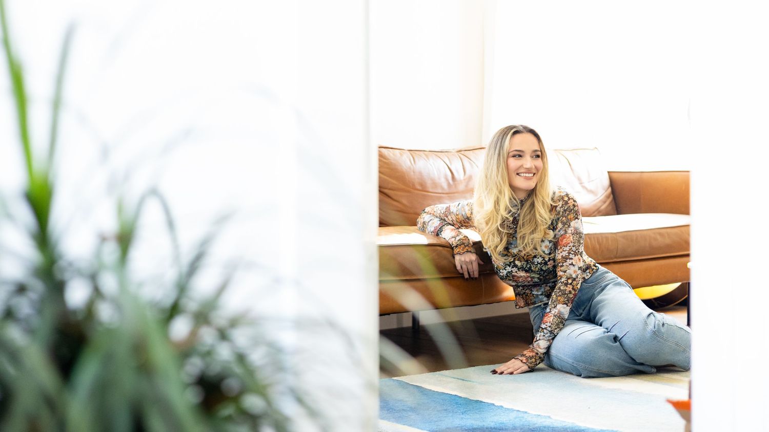 Blonde woman in a patterned top and jeans smiles while sitting on the floor in front of a leather sofa, framed by a leafy plant.