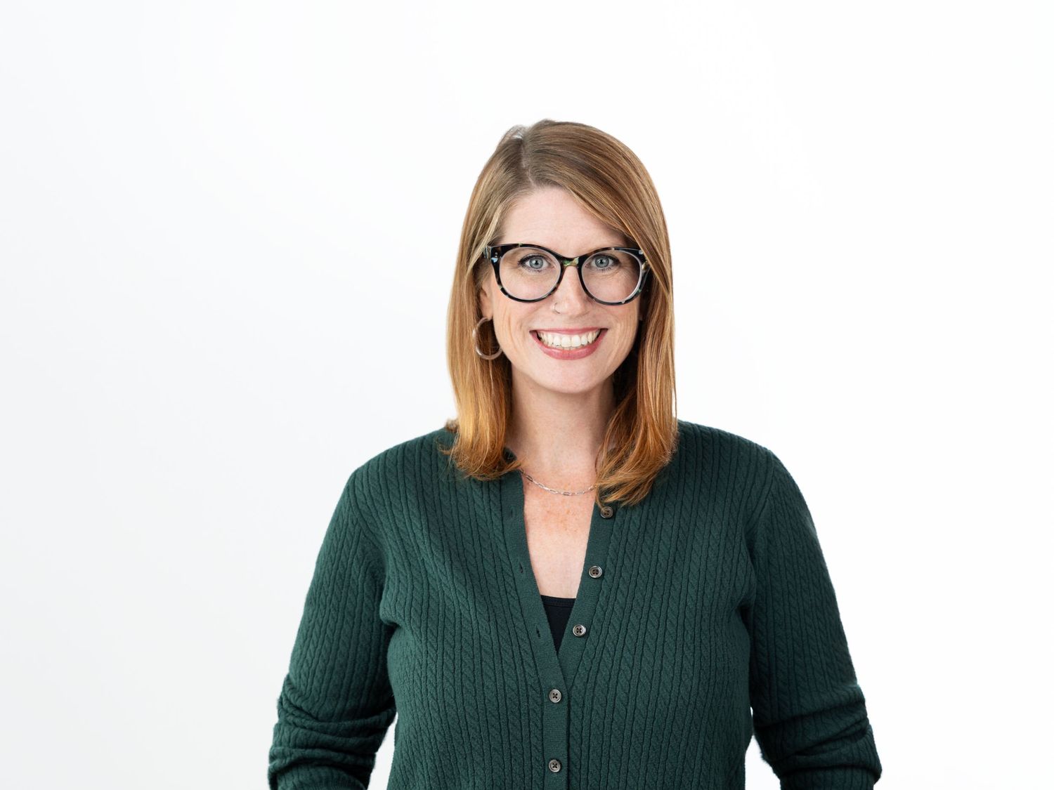 Smiling woman with shoulder-length auburn hair and black-framed glasses wearing a dark green cardigan in a bright studio.