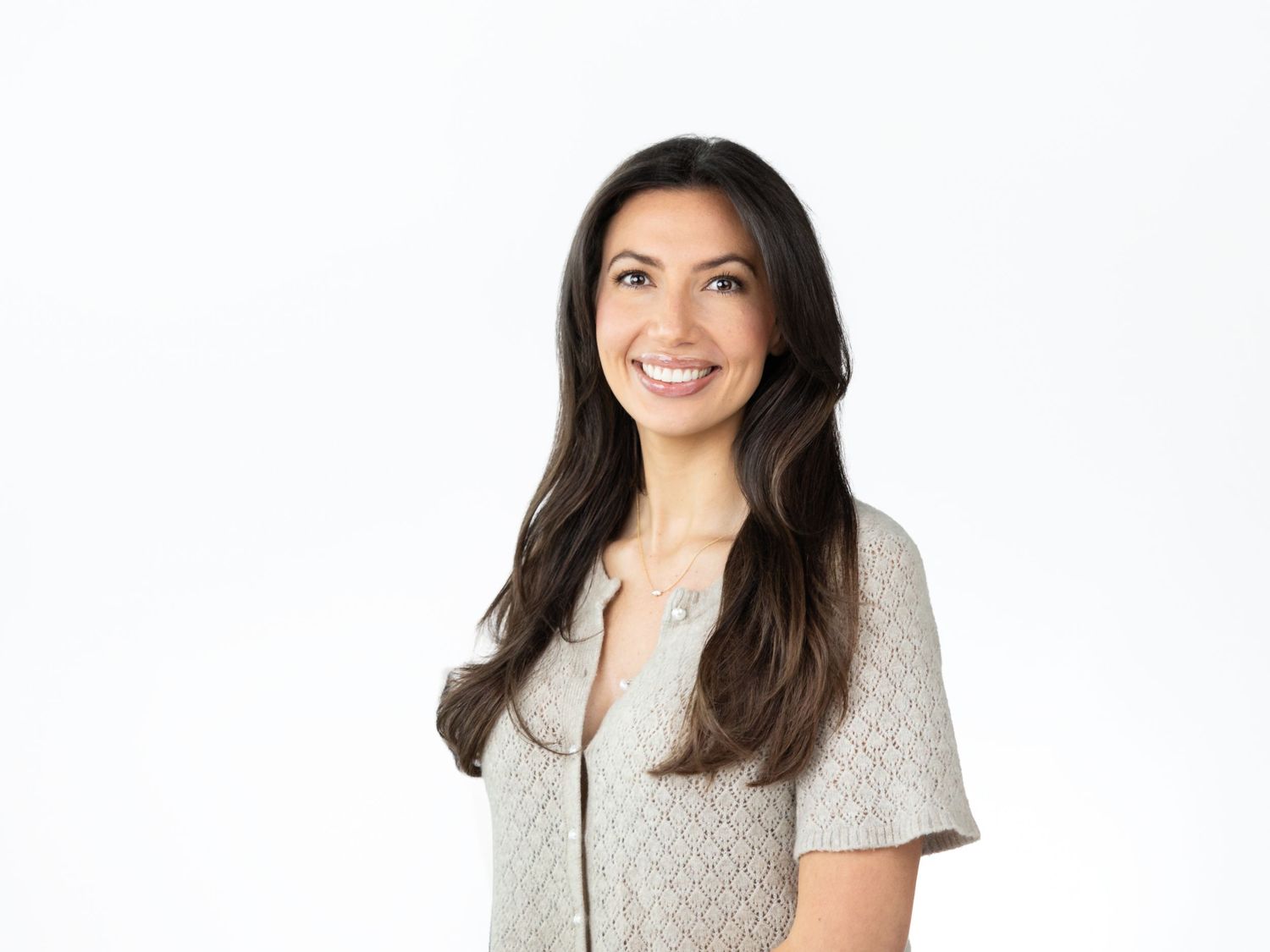 Smiling woman with long dark hair wears a light textured short-sleeve top in a professional headshot against a white background.