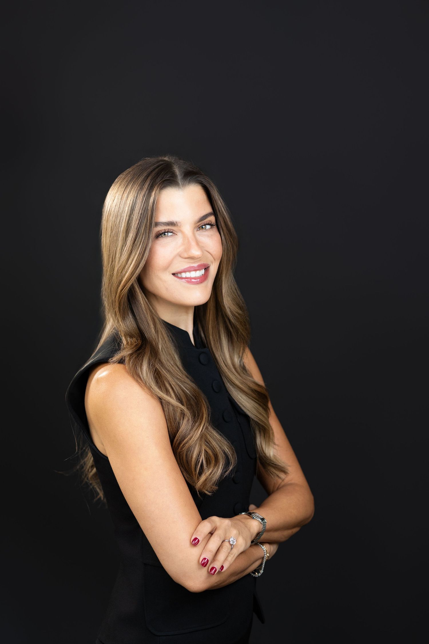 Professional portrait of a smiling woman with long highlighted brown hair in a black sleeveless top against a black background.