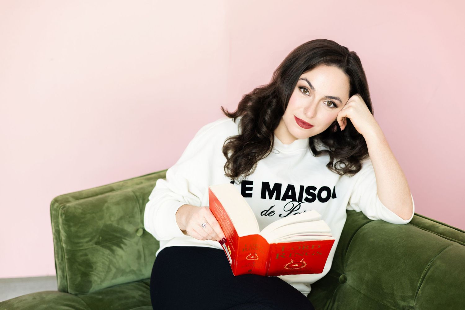 Woman in a Le Maison de Paris sweatshirt sits on a green velvet sofa holding an open red book near a pink wall.