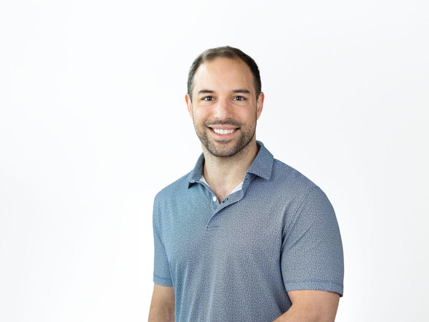 Smiling bearded man with short dark hair wearing a blue patterned polo shirt posing against a white background.